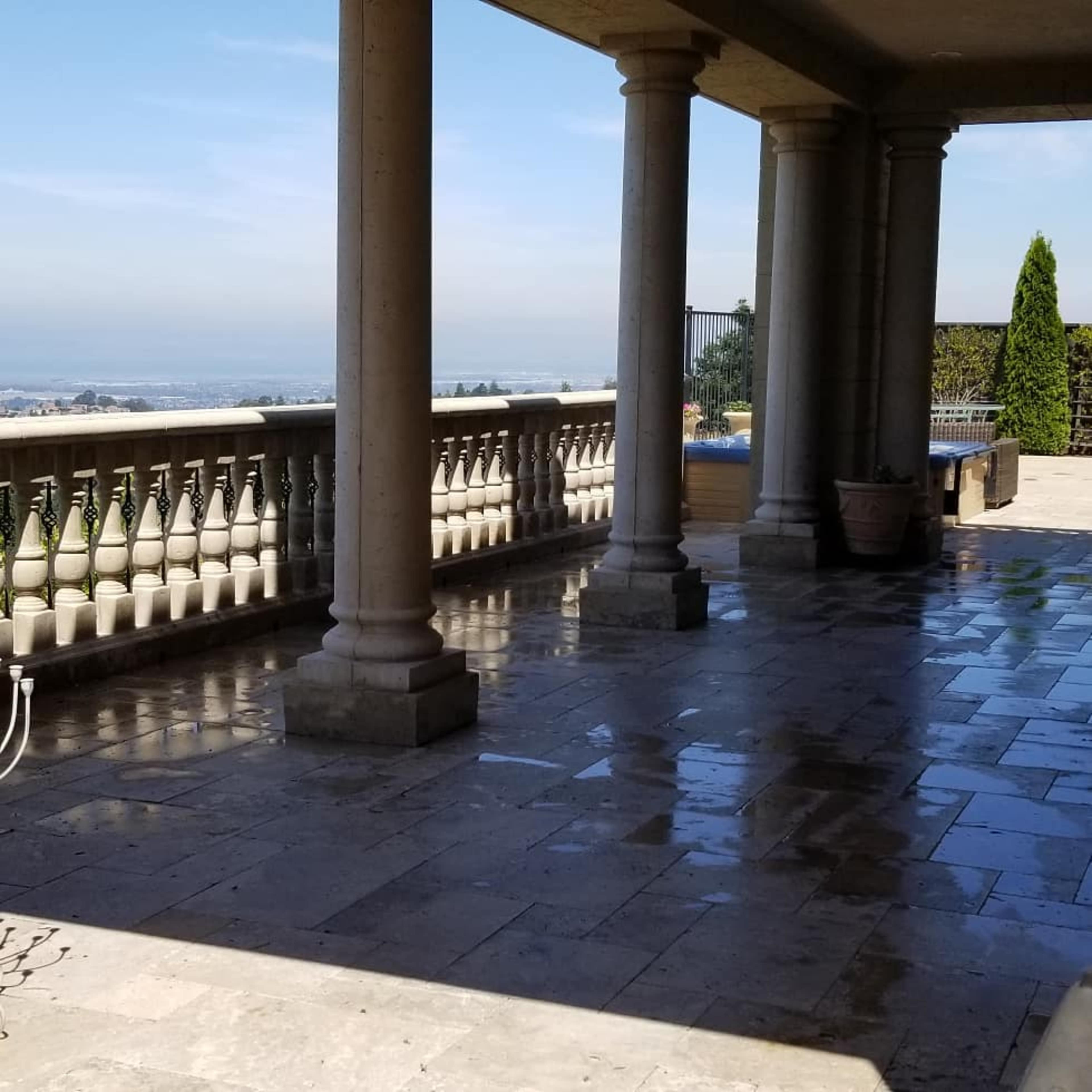 The image shows a spacious stone terrace with columns and a view of the distant landscape under a clear sky.