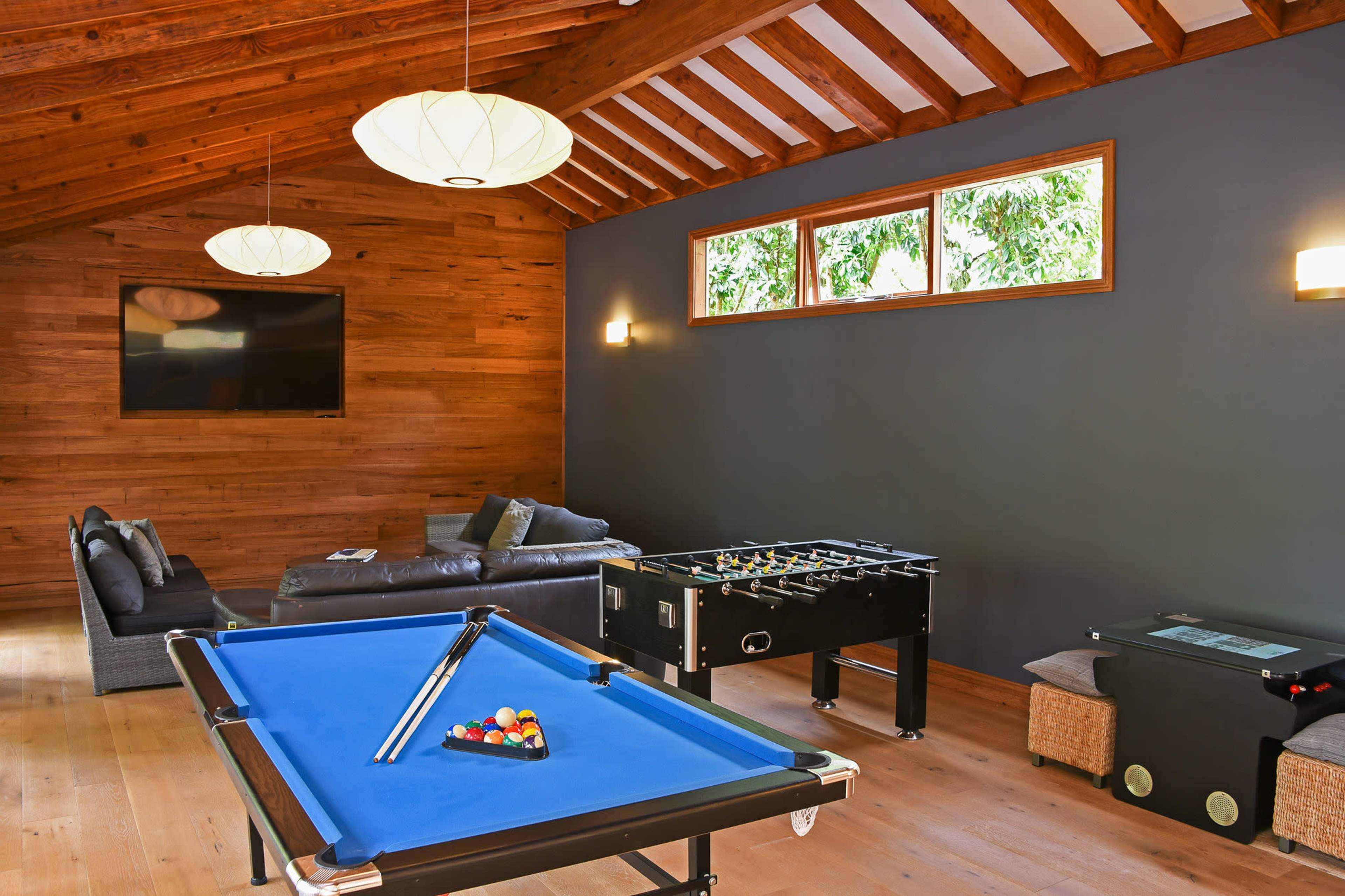 A game room features a blue pool table, a foosball table, and a sectional sofa under wooden beams with large windows allowing natural light.