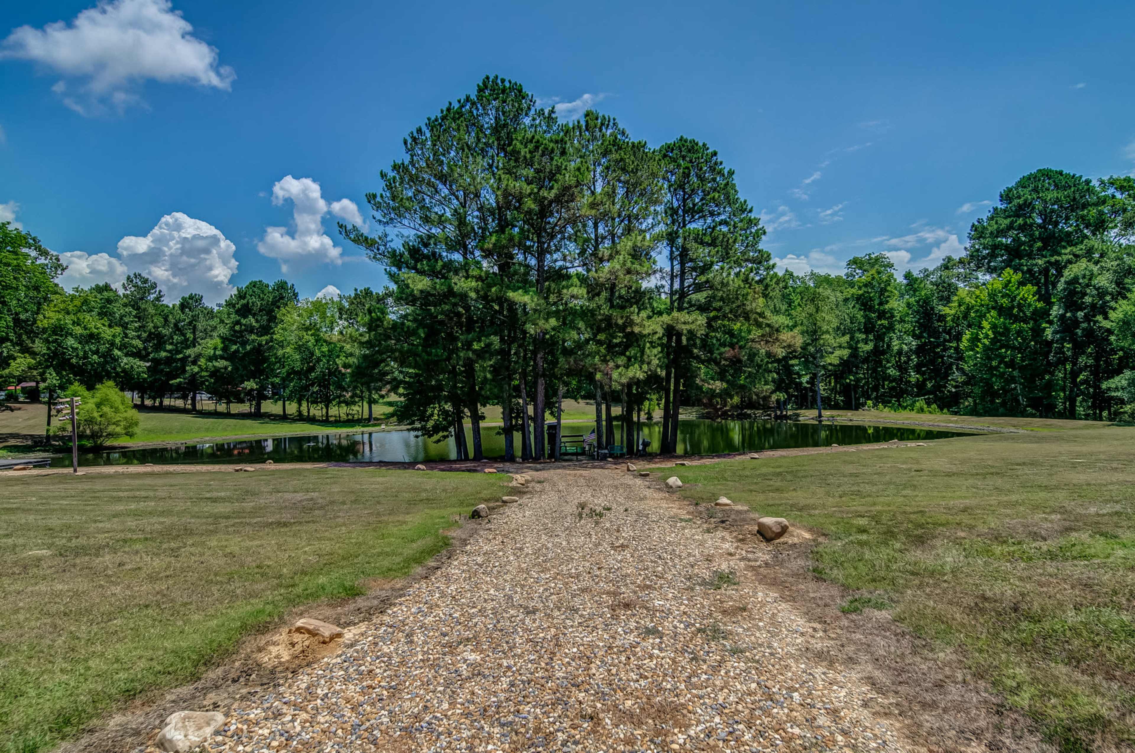 A gravel path leads to a small pond surrounded by trees in a green landscape.