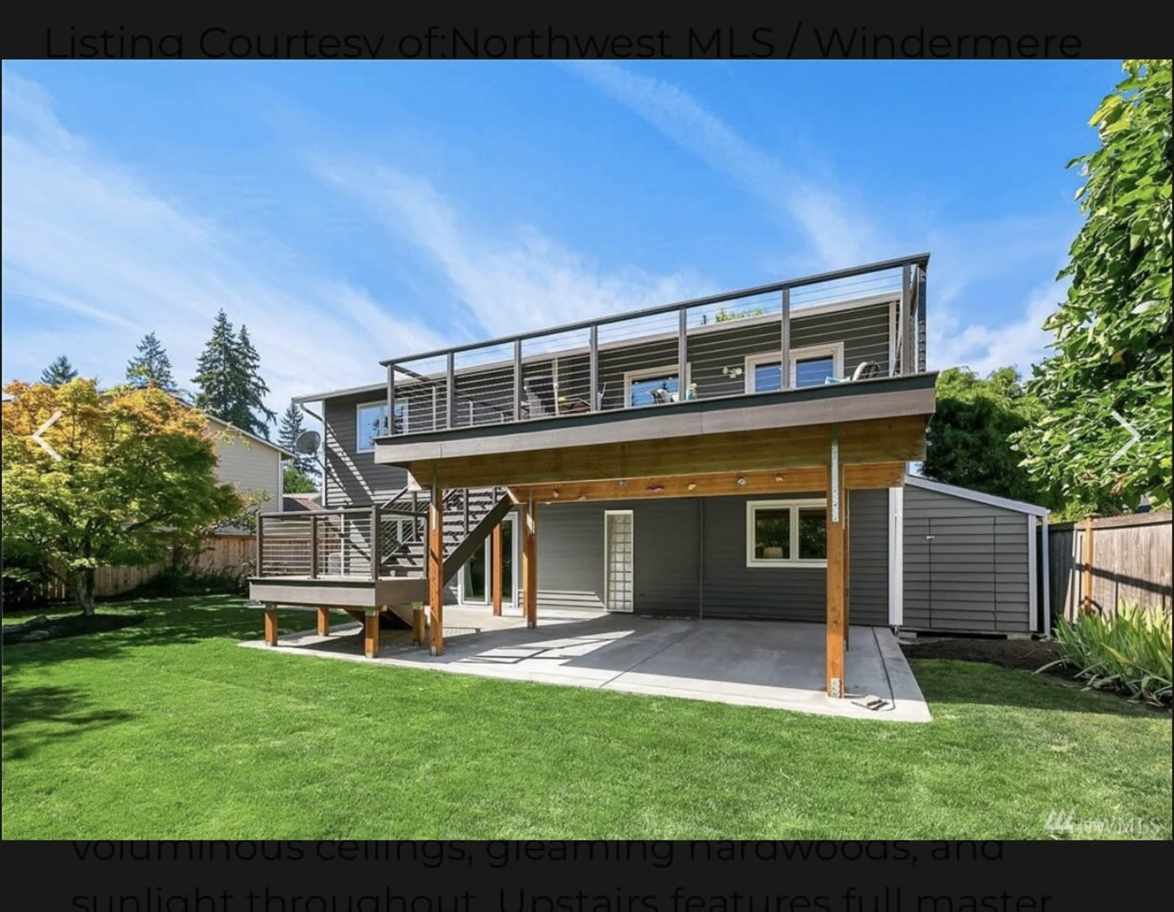 A modern two-story house with a spacious deck overlooking a green lawn in a residential area.