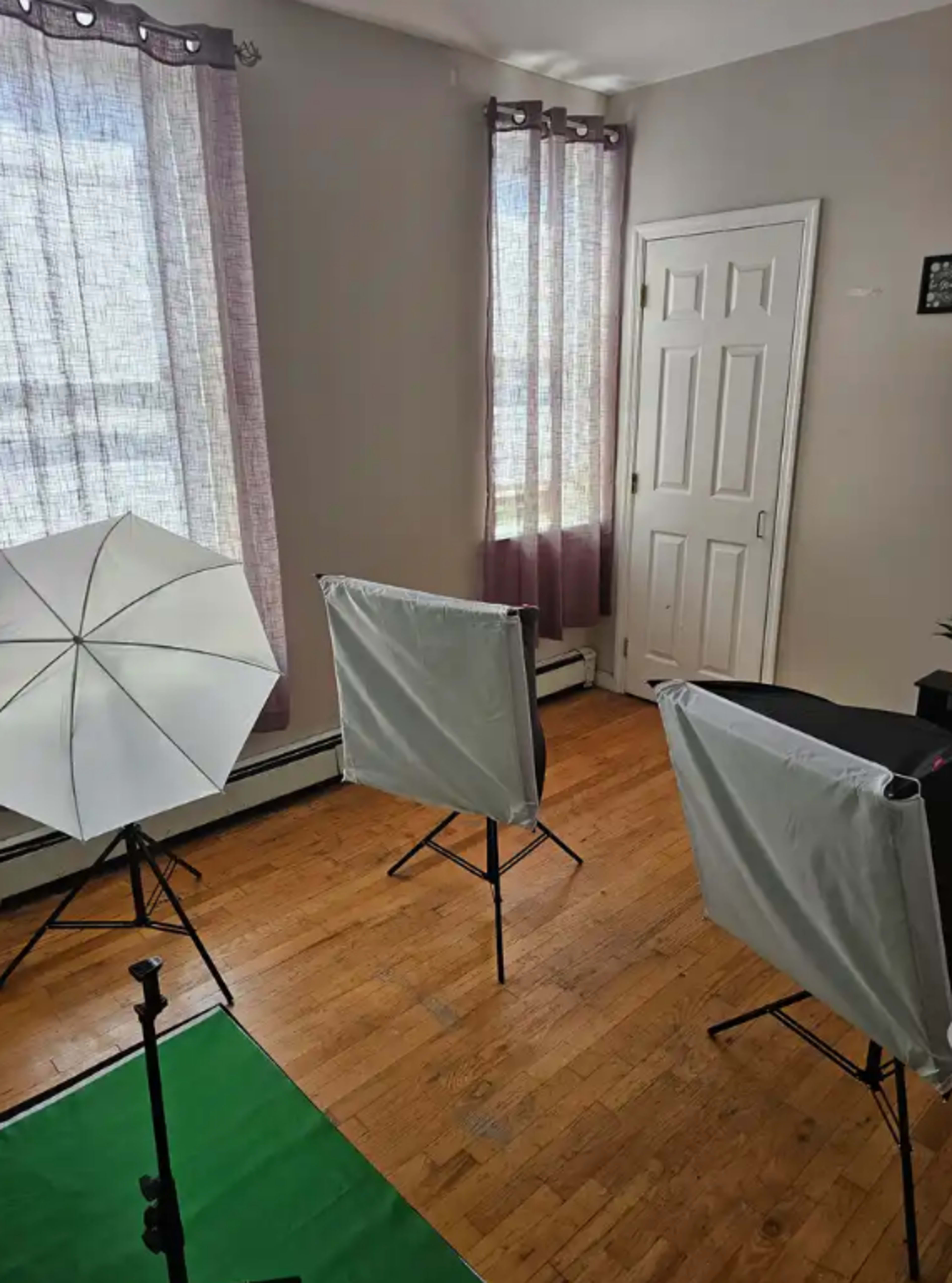 The image shows a bright room with wooden flooring, two softbox light stands, and a green backdrop set up for photography or video production.