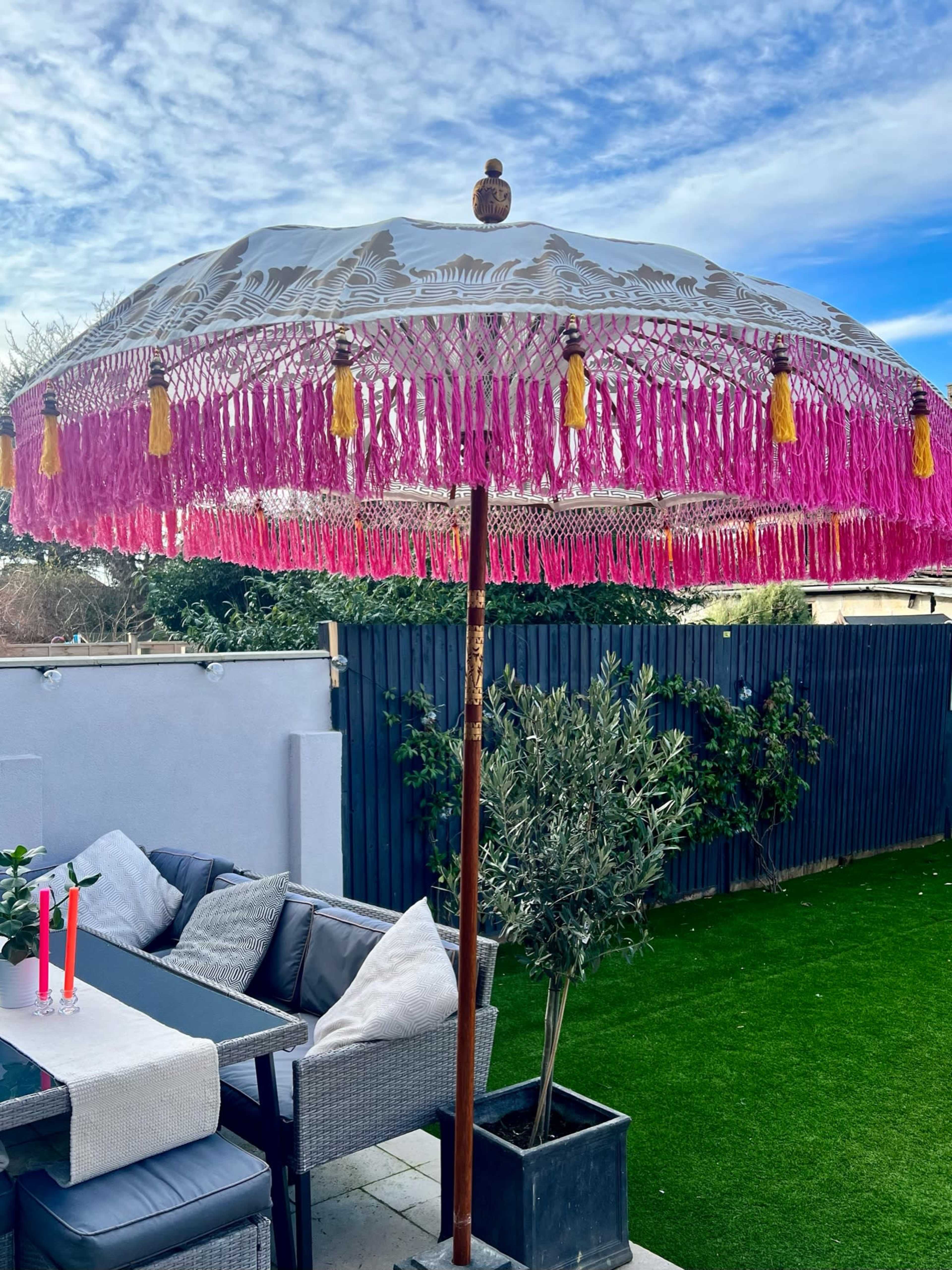 A large decorative umbrella with pink fringe and tassels stands in a garden area beside a seating arrangement.
