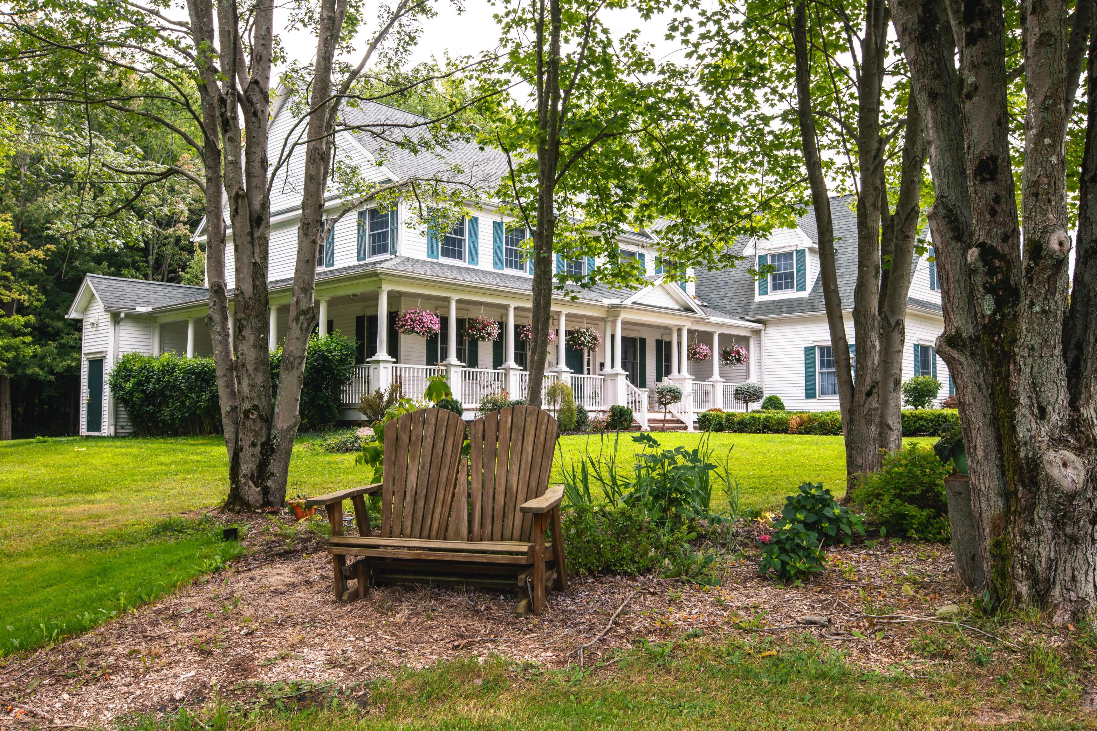 A wooden adirondack chair sits in a landscaped yard in front of a large two-story house surrounded by trees.
