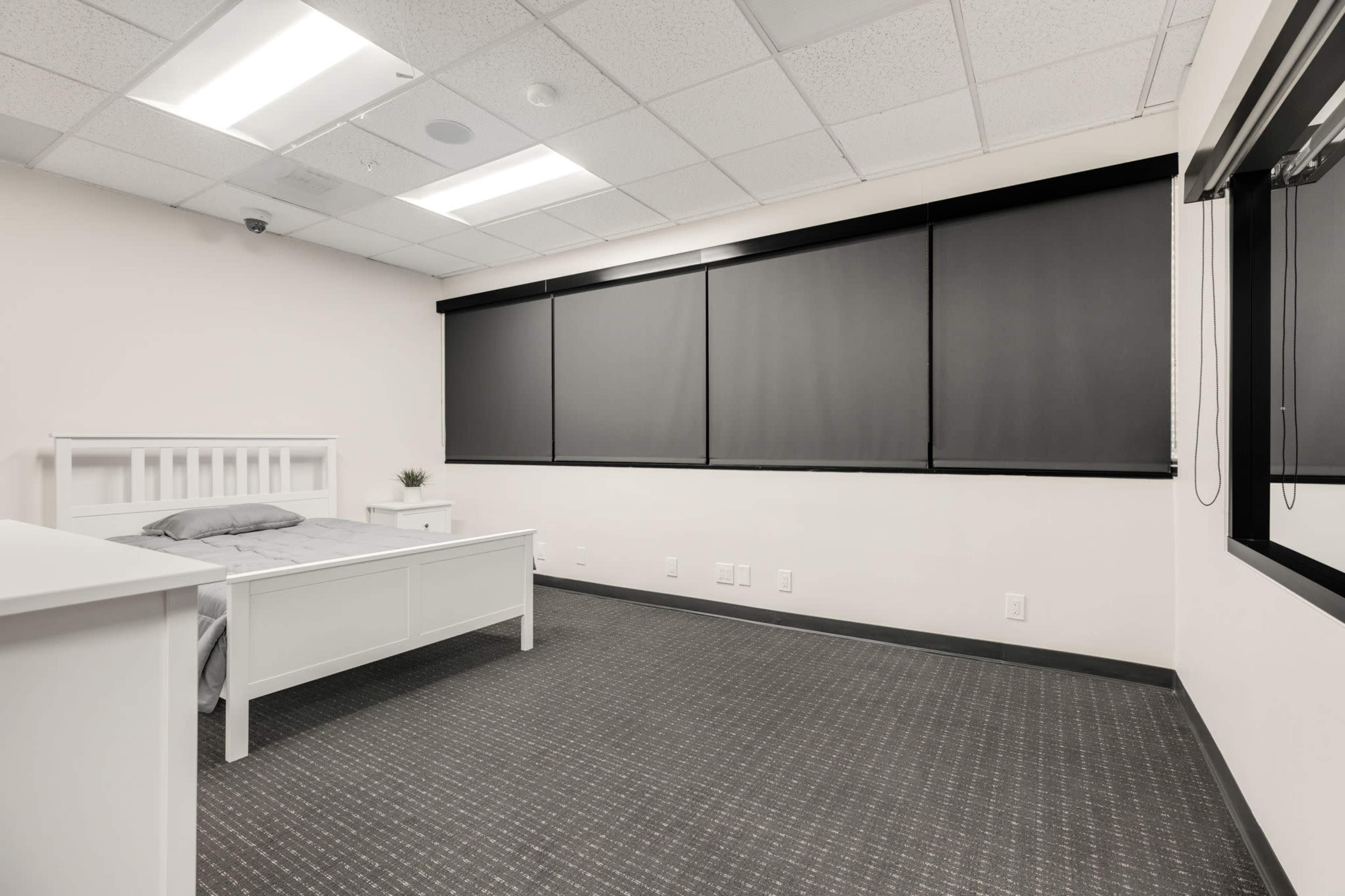 The image depicts a minimalist bedroom featuring a white bed frame, grey bedding, and large dark window coverings on a light-colored wall.