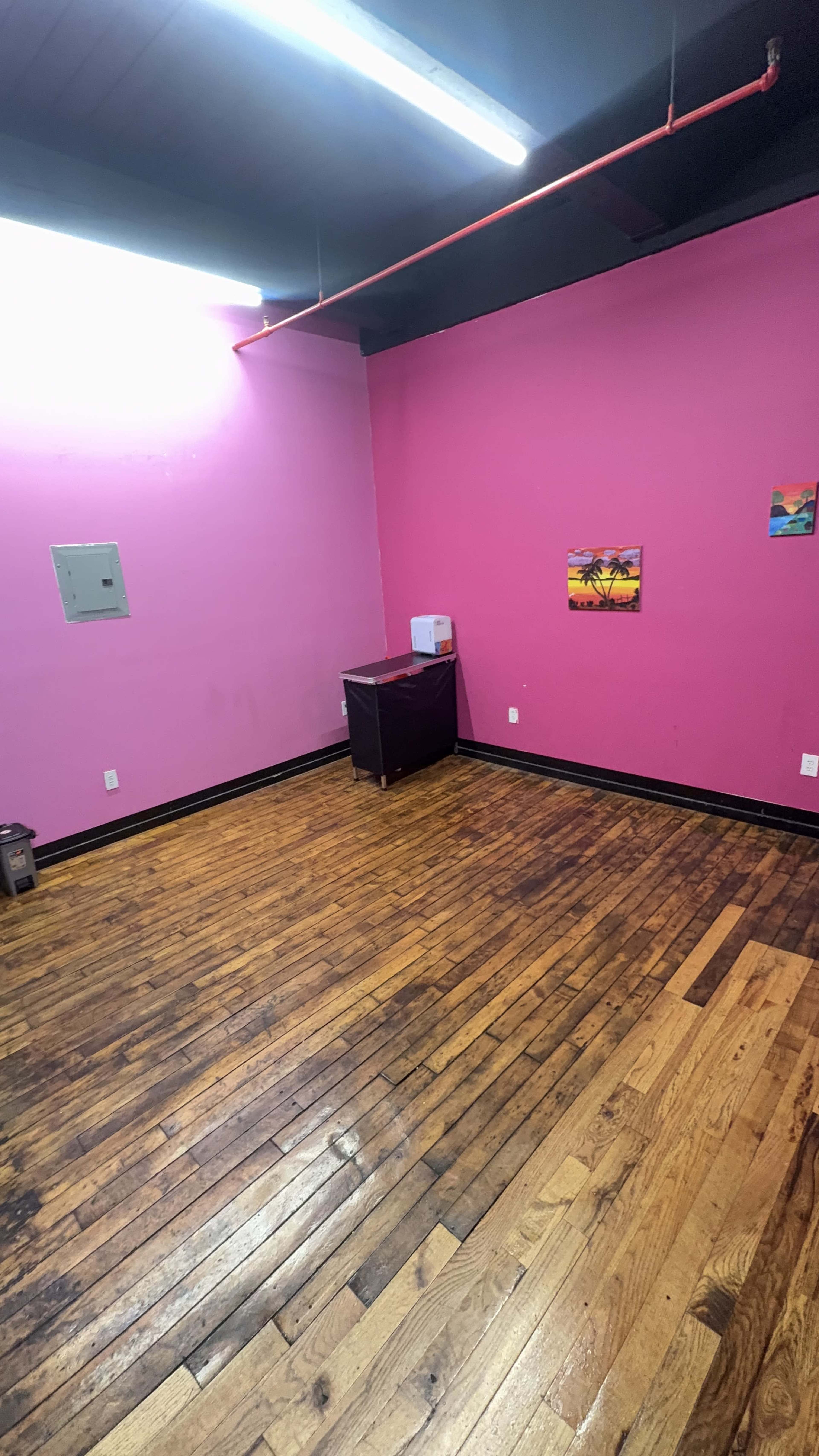 The image shows an empty room with pink walls, wooden flooring, and a small dark cabinet in one corner.