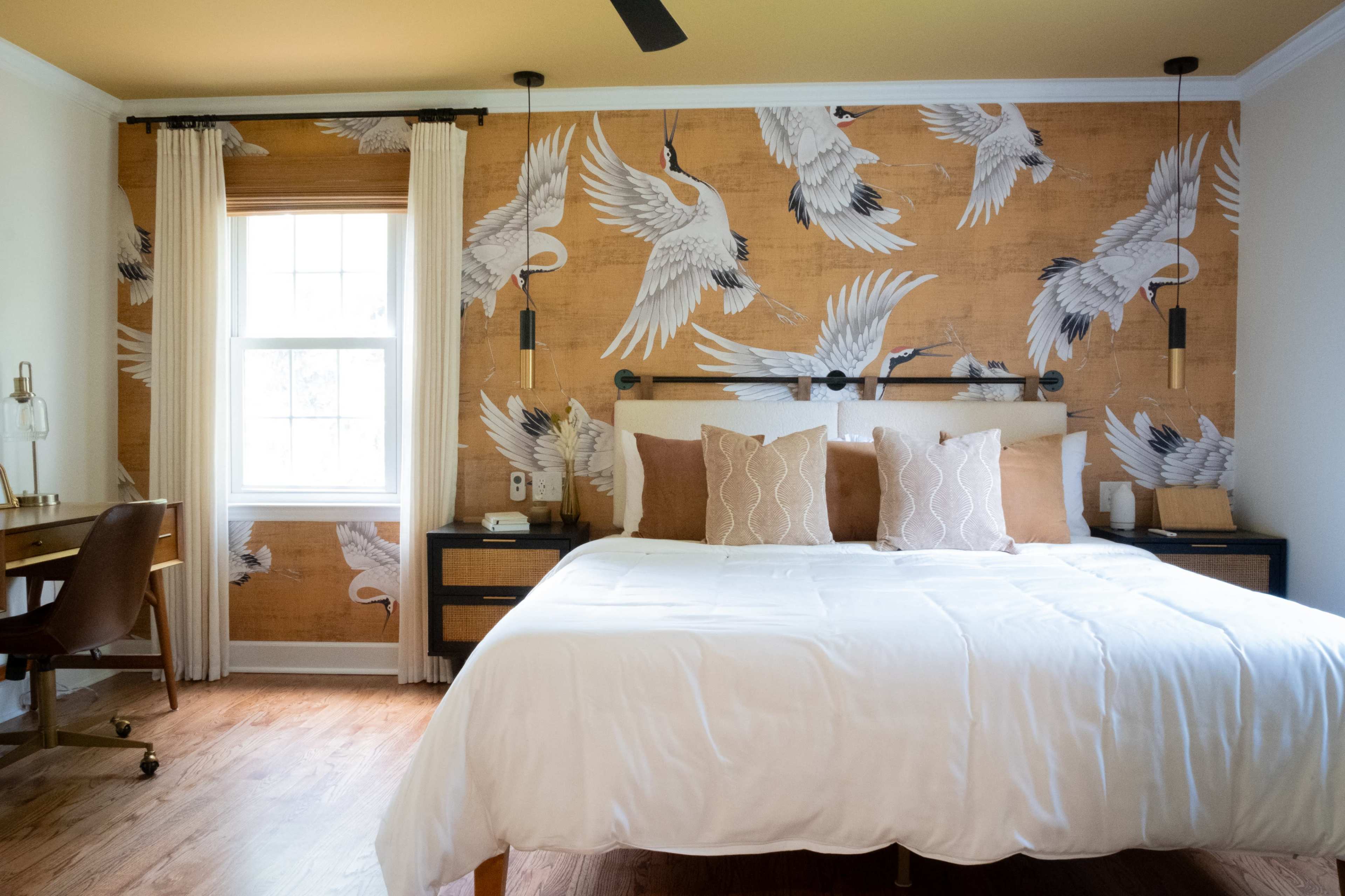 The bedroom features a large bed with white linens, flanked by pillows, and a wall adorned with a crane-patterned wallpaper, complemented by a window and a small desk area.
