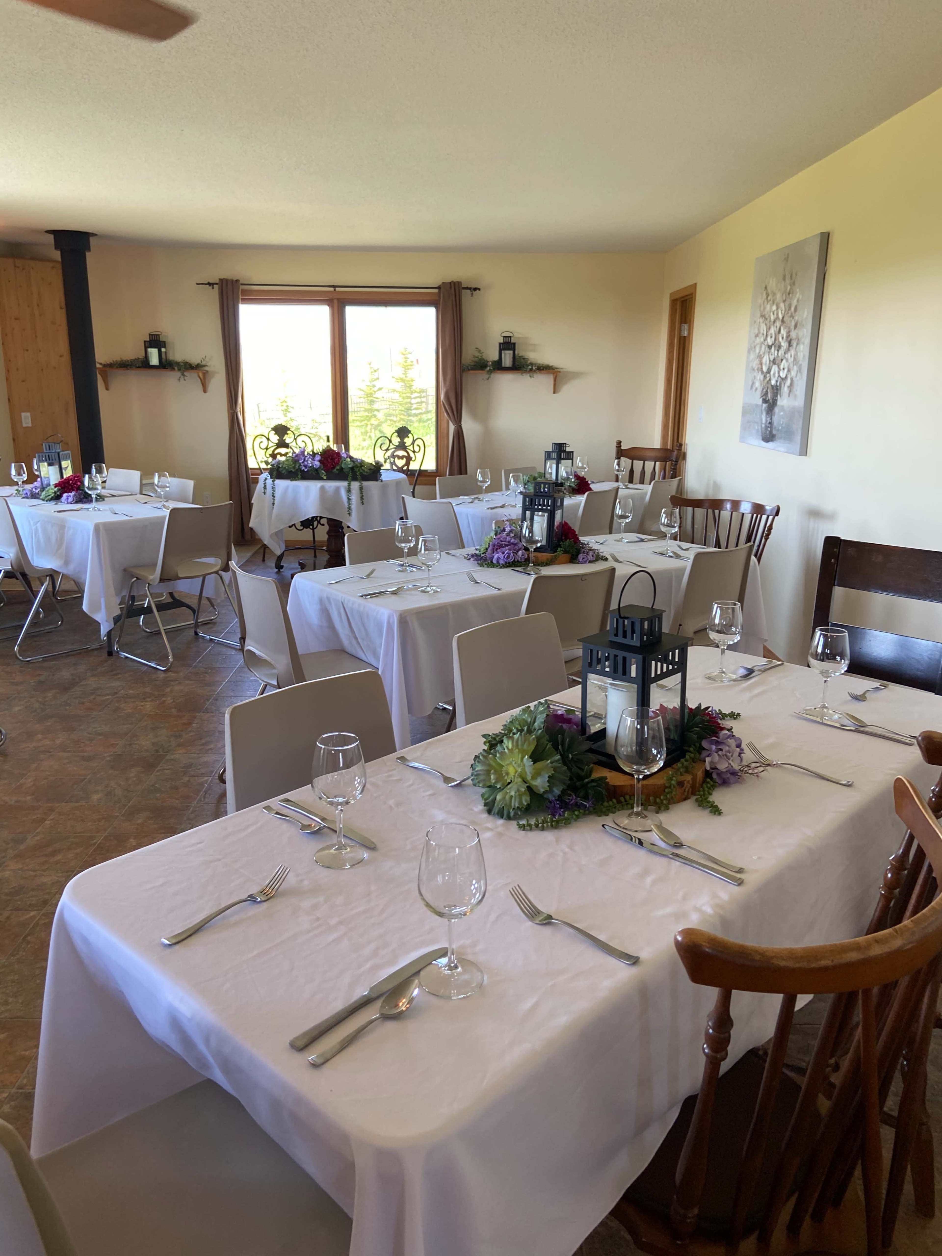 The image shows a dining room with several tables set with white linens, glassware, and floral decorations, ready for a gathering or event.