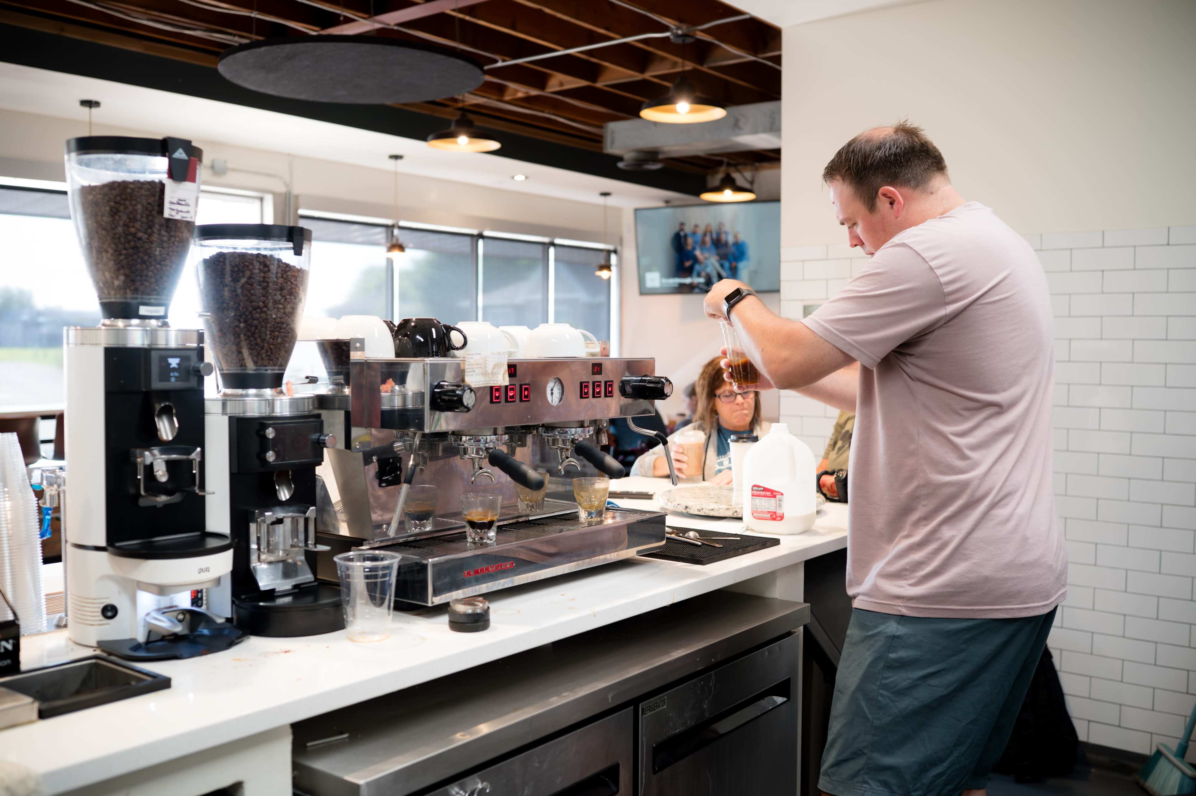A barista prepares a coffee drink at a cafe while a customer waits at the counter.