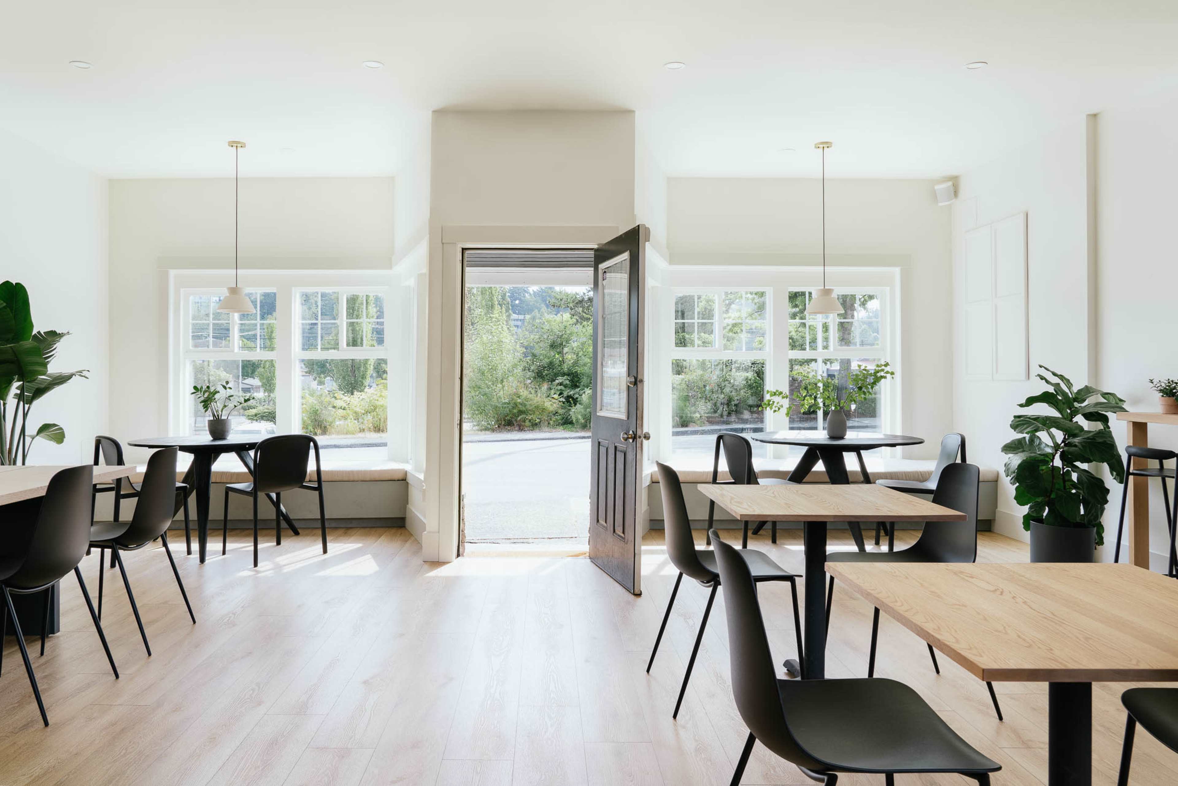 A bright, minimalist café features wooden tables, black chairs, and large windows with potted plants, opening to an outdoor view.
