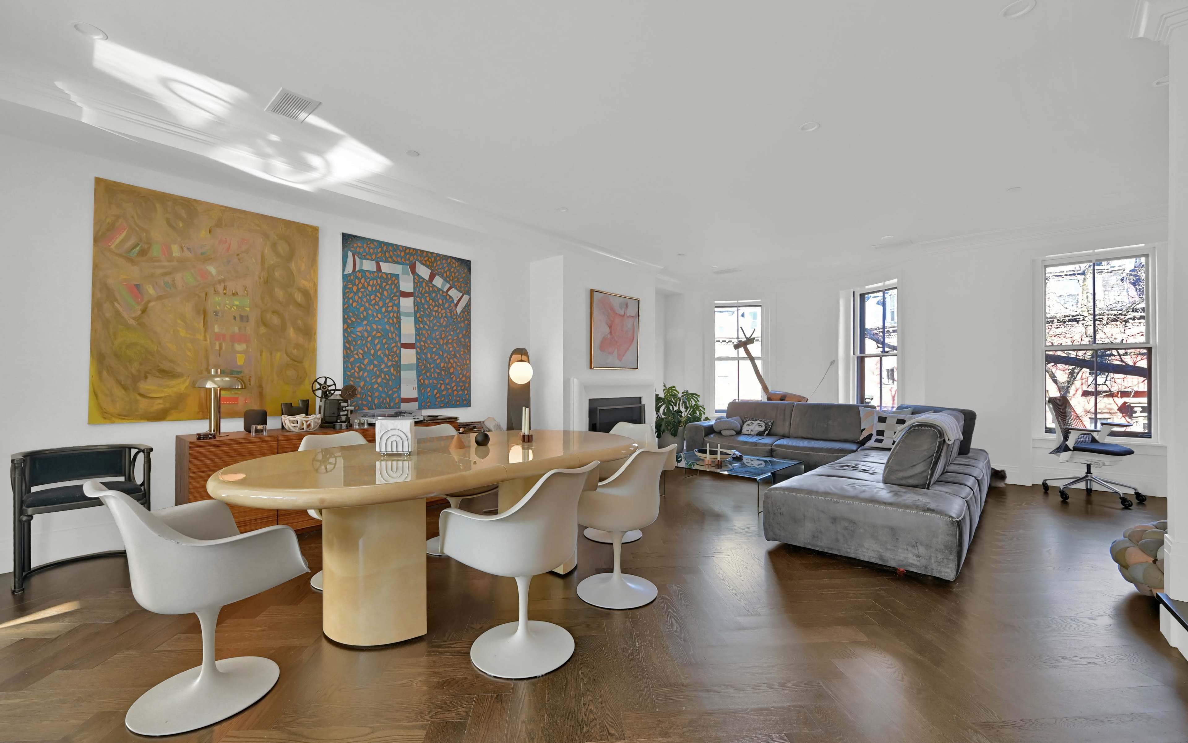 A modern living room with a round dining table surrounded by white chairs, two large paintings on the walls, and a gray sofa facing a fireplace and windows.