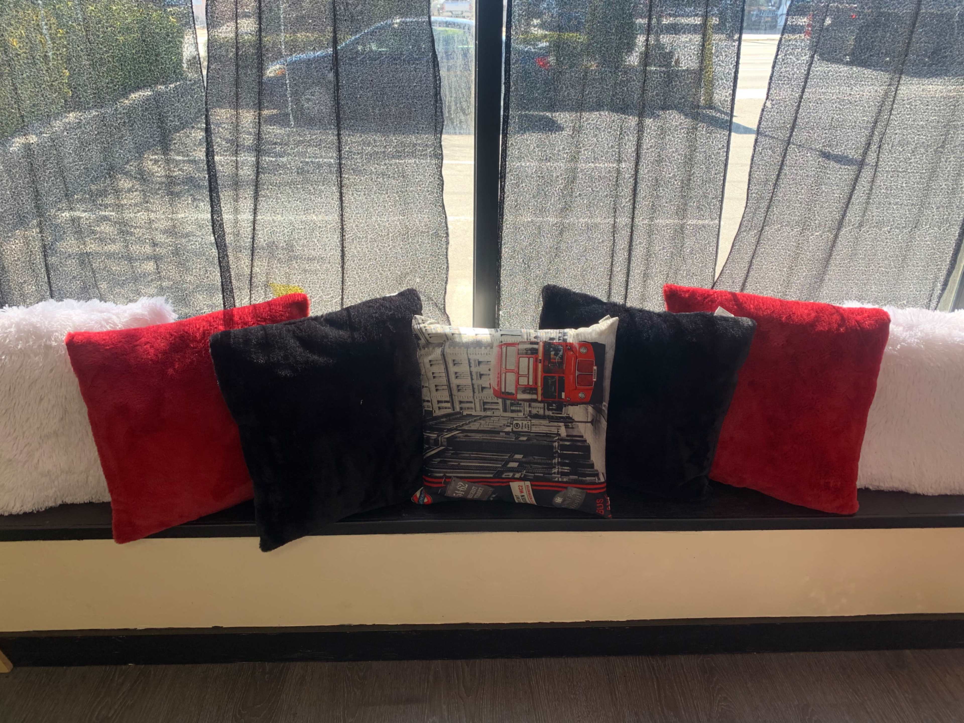 A row of decorative pillows in red, black, and white is arranged on a windowsill with sheer curtains behind them.