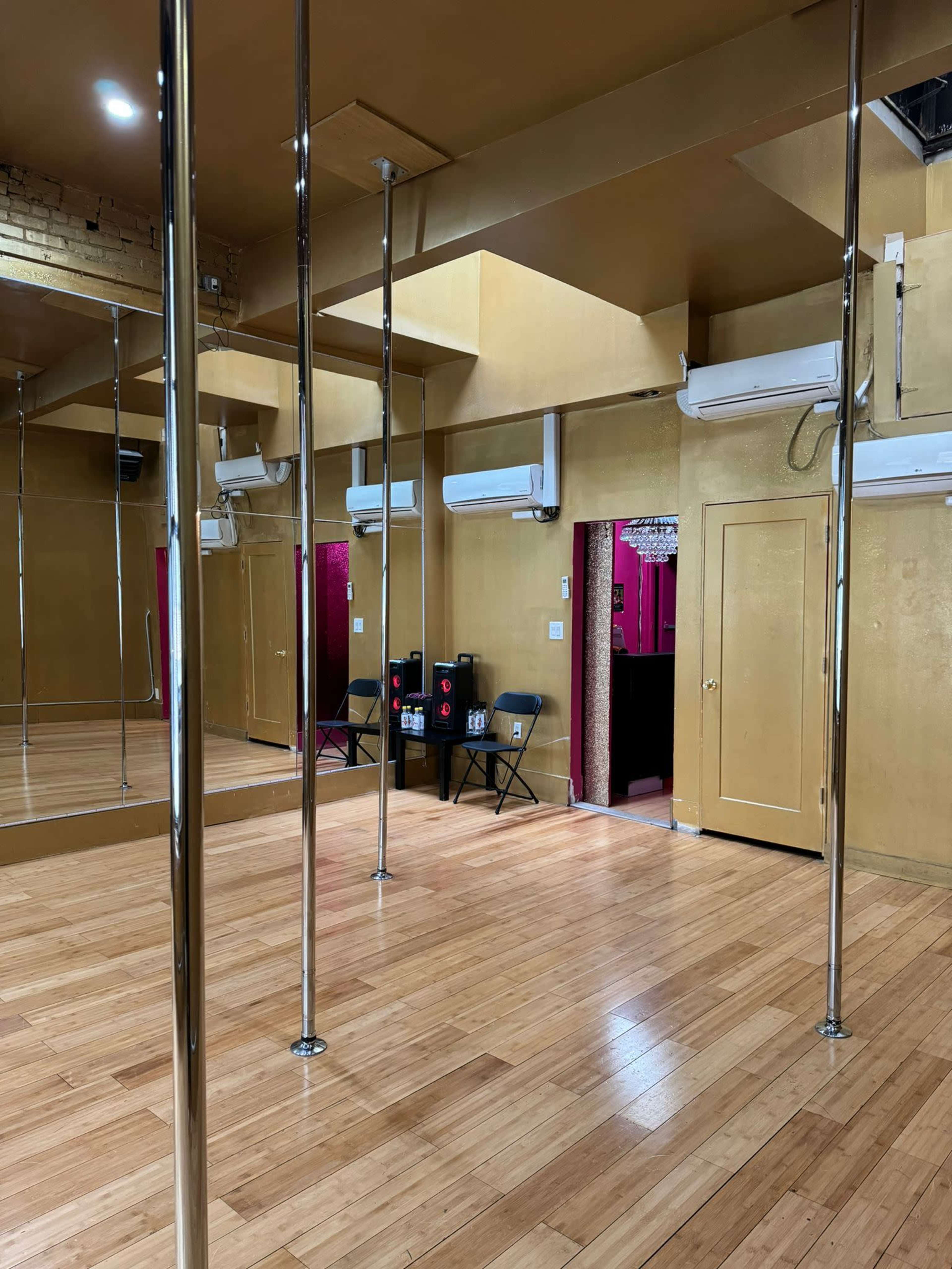 A spacious dance studio with a polished wooden floor, mirrors, and metallic poles, featuring chairs stacked near the entrance.