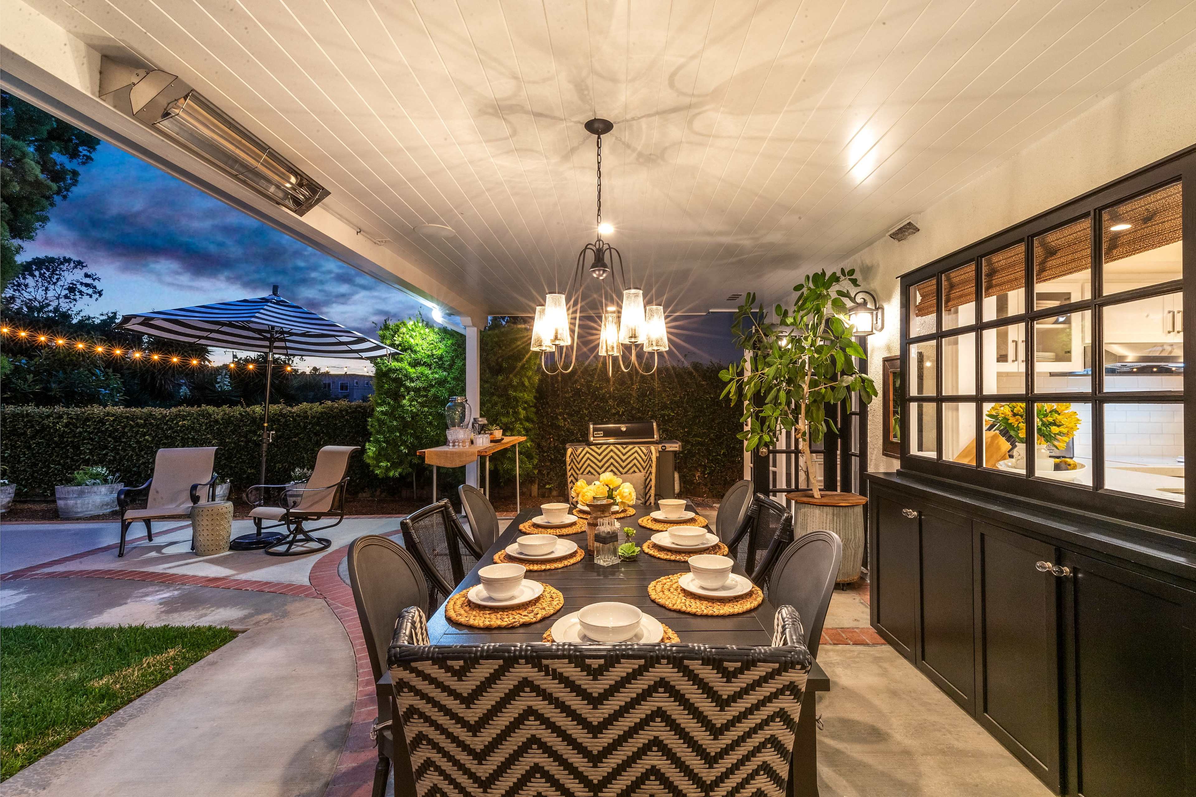 A spacious outdoor dining area with a table set for dinner, surrounded by greenery and string lights under a covered patio.