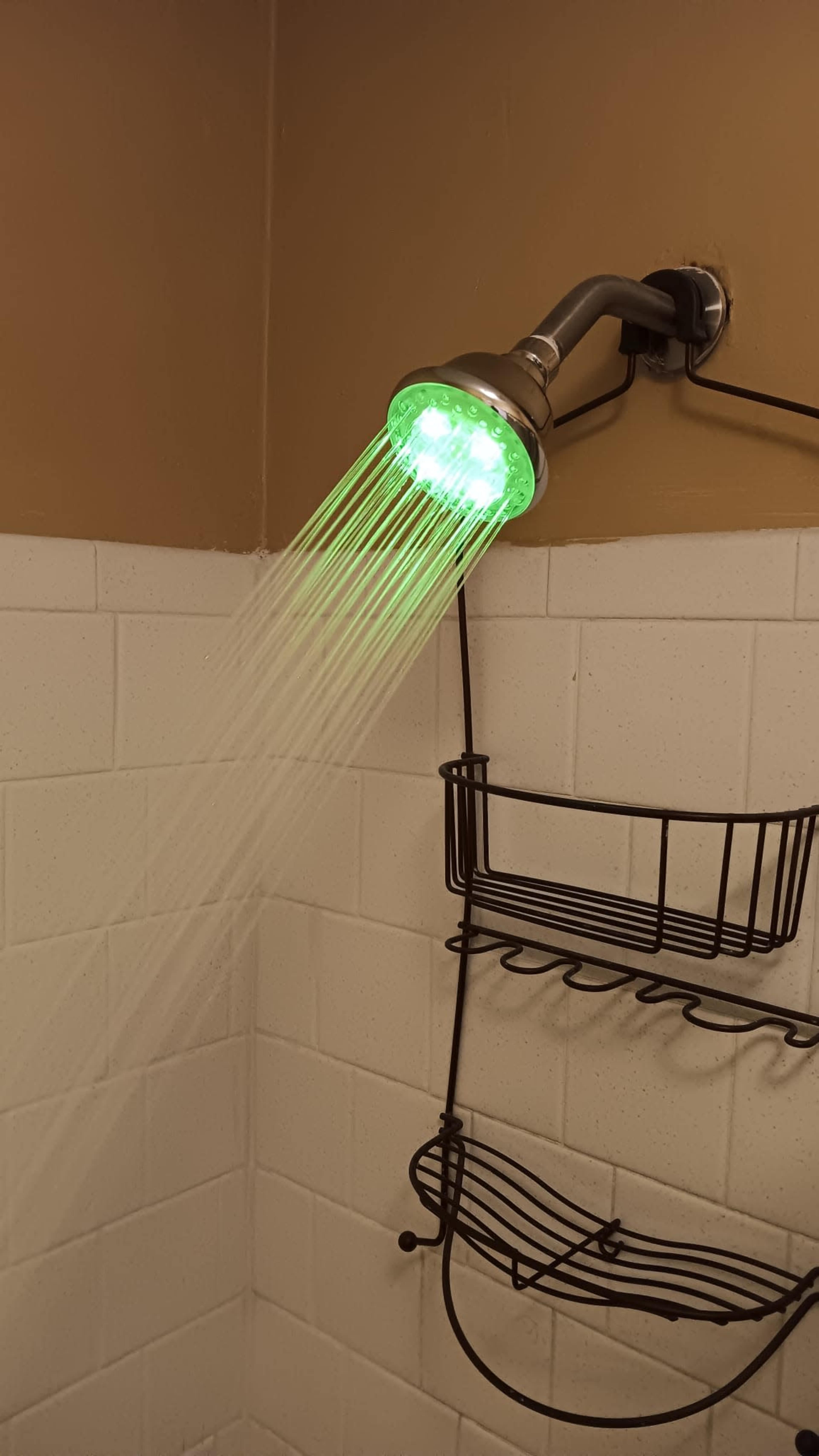 A showerhead emits green light while water flows from it, with a metal shower caddy hanging nearby.