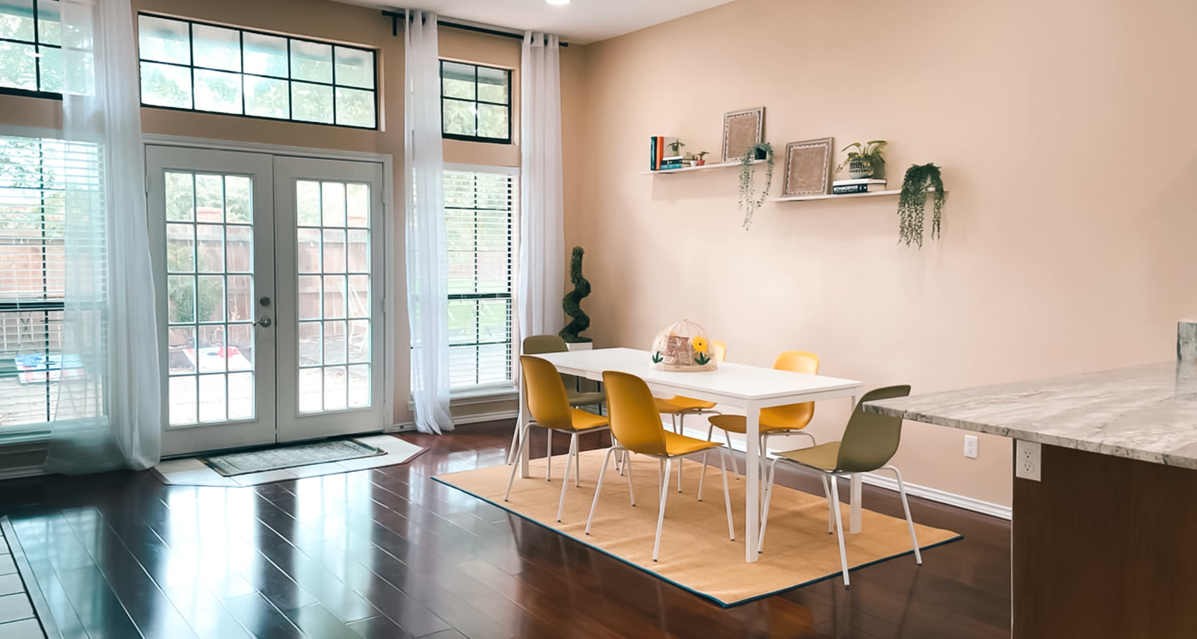 A modern dining area features a white table with colorful chairs surrounded by large windows and a sliding glass door leading to an outdoor space.