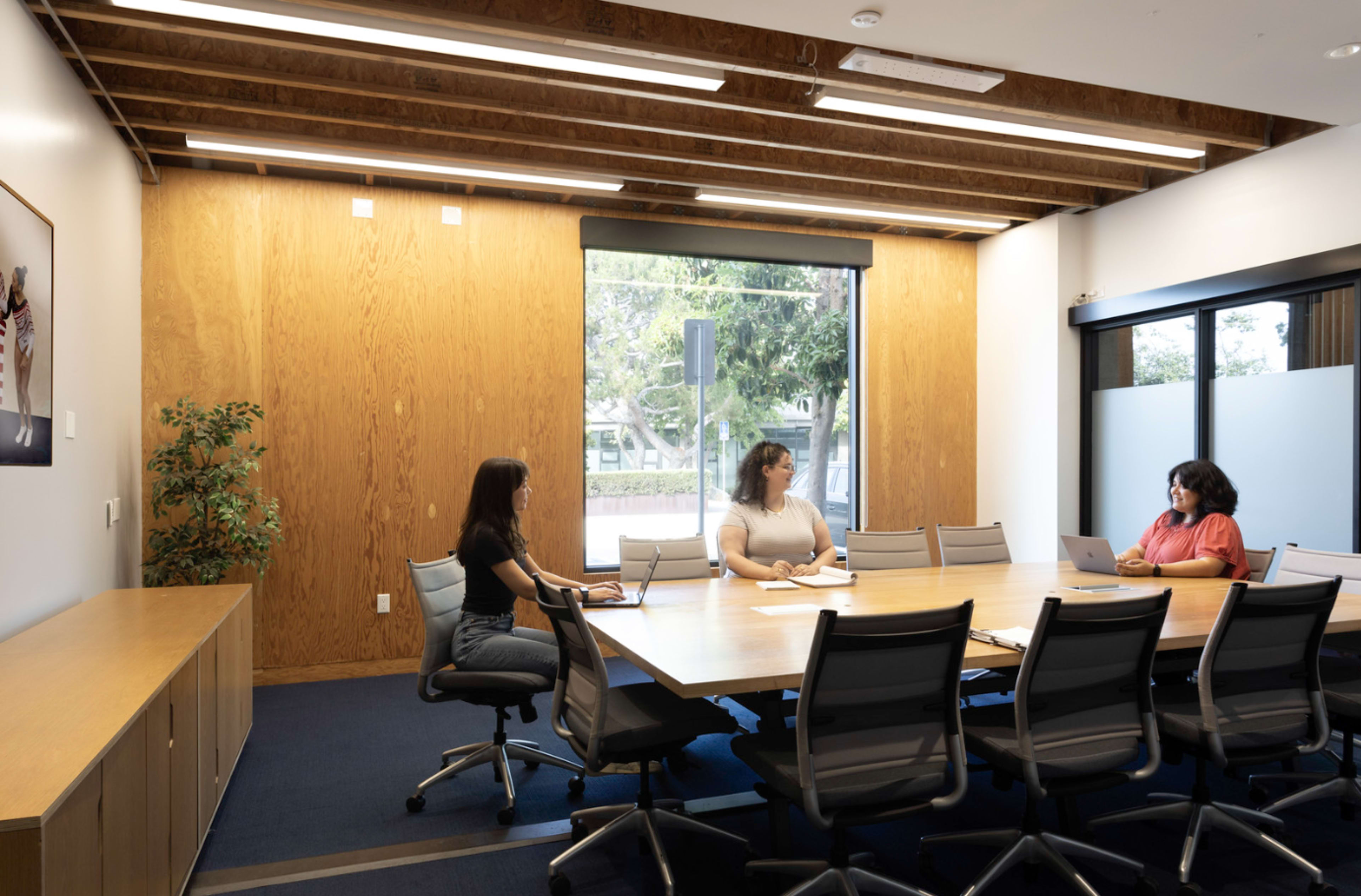 Three women sit at a large conference table in a modern meeting room with a wooden wall and a window.