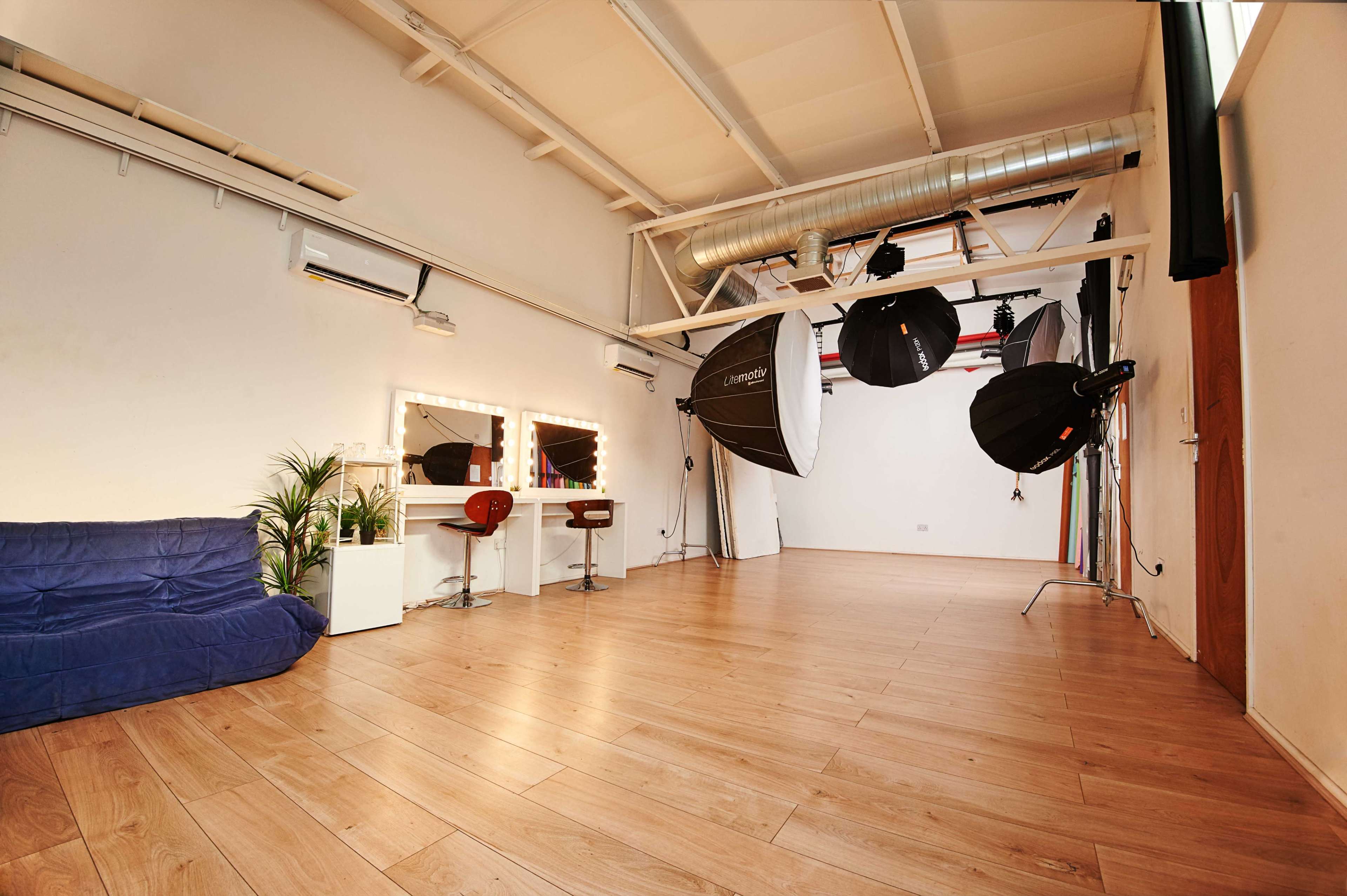 A spacious photography studio featuring a couch, a makeup station with lights, and several photography umbrellas suspended from the ceiling.