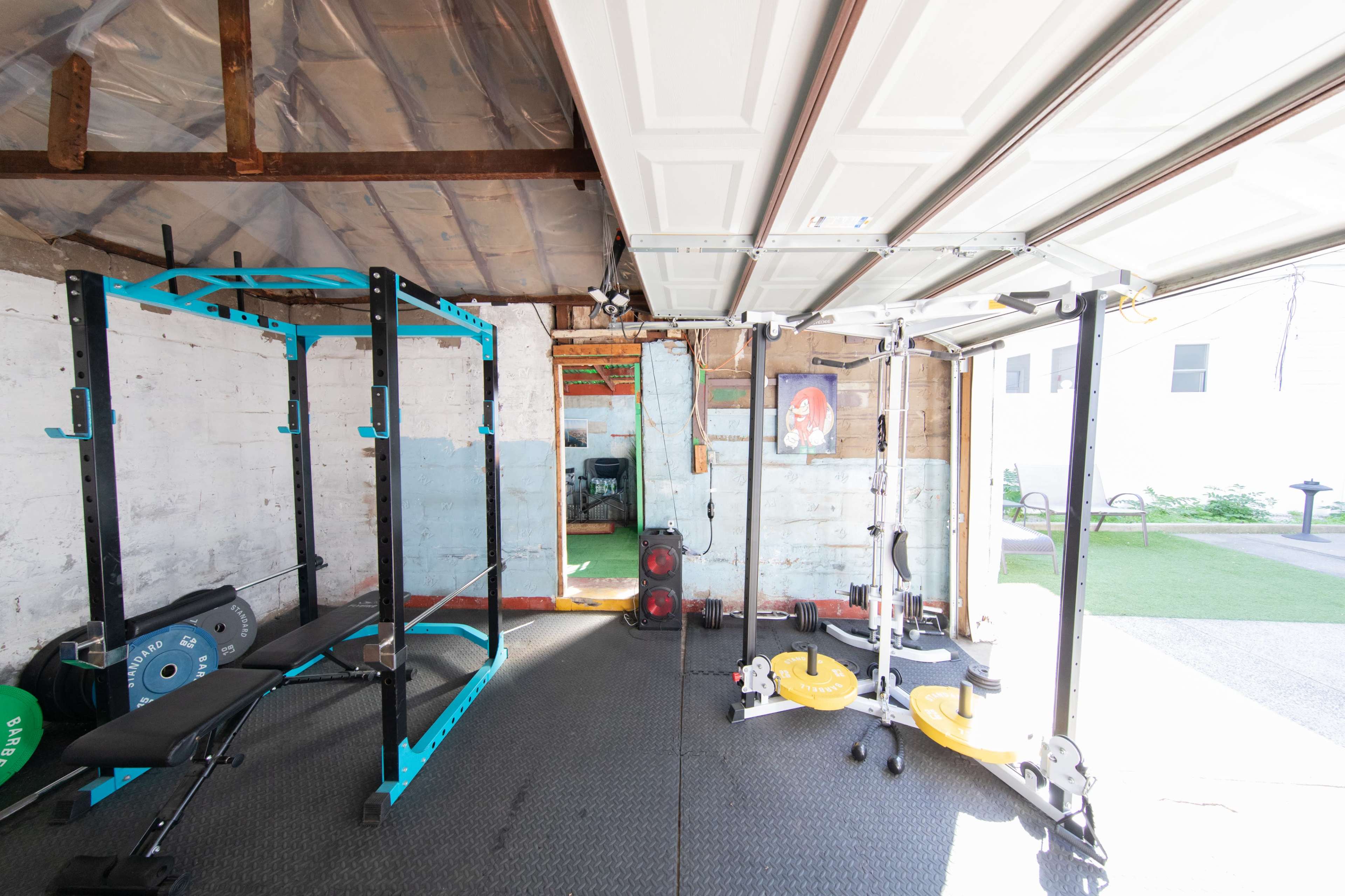 The image shows a partially finished home gym with workout equipment including a squat rack, a bench, and a cable machine, set inside a garage space.