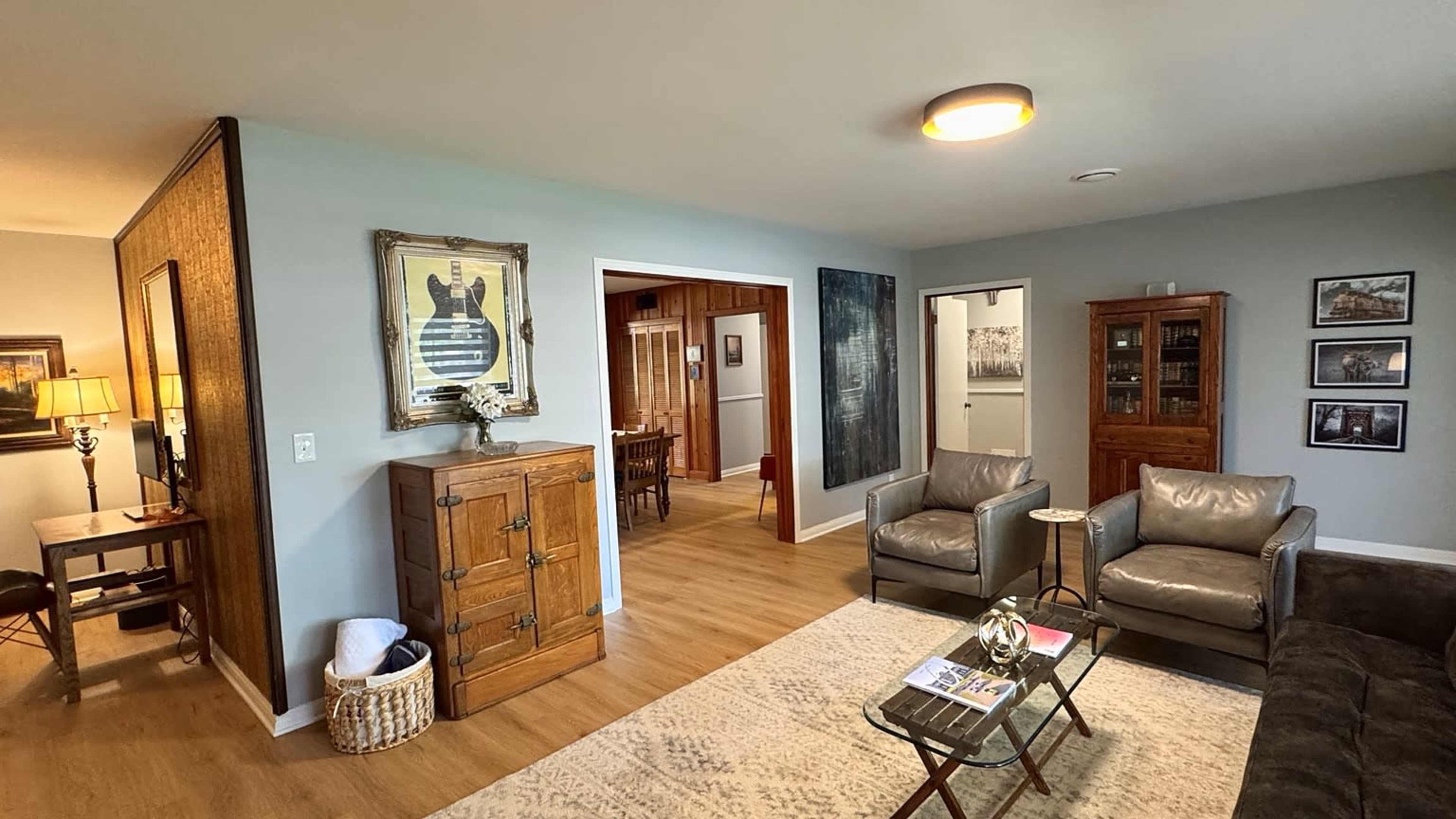 The image shows a cozy living room featuring two leather armchairs, a coffee table, and a wooden cabinet, with a view into a dining area in the background.