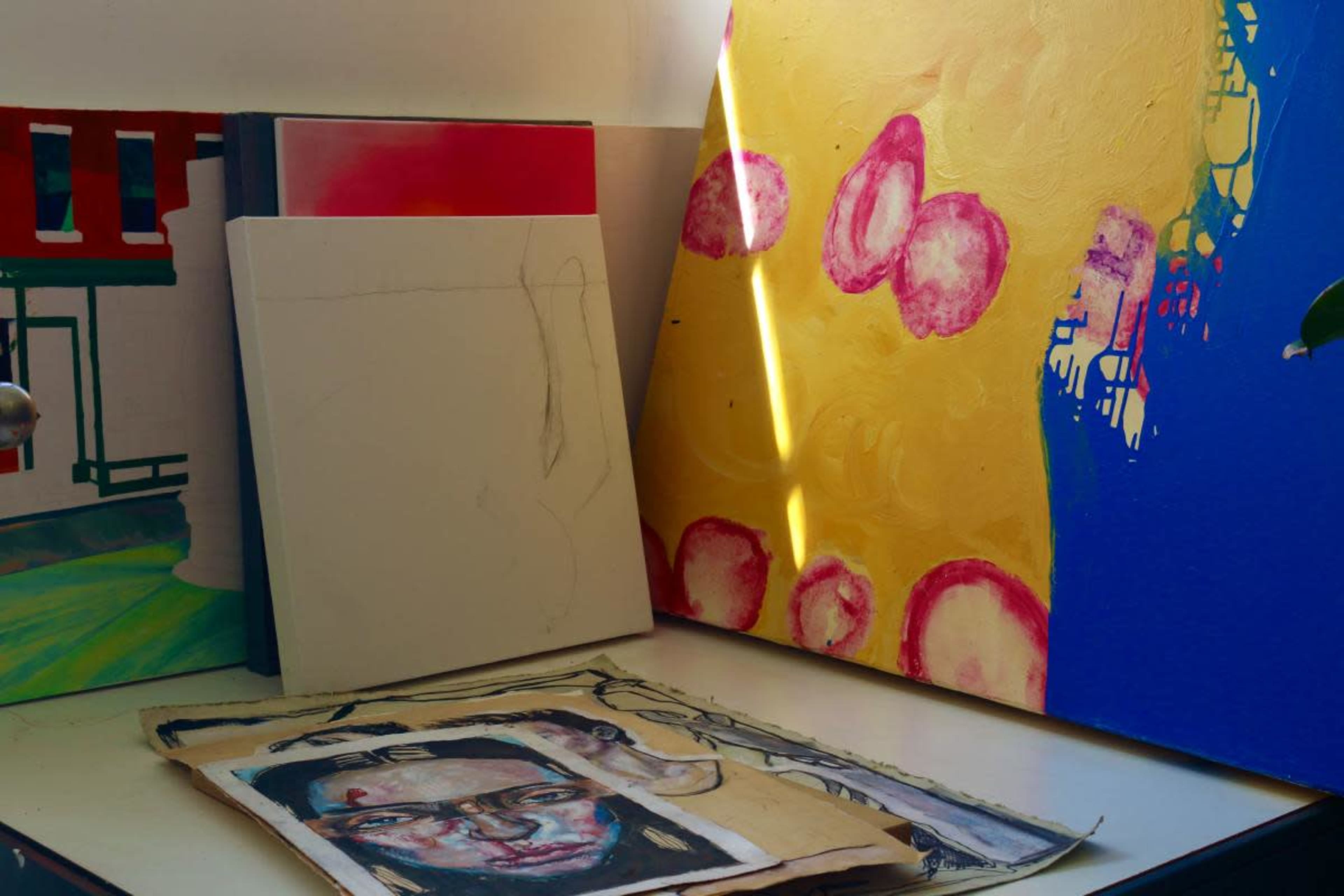 The image shows a collection of artworks and canvases arranged on a table, including a partially finished painting and a colorful abstract piece.