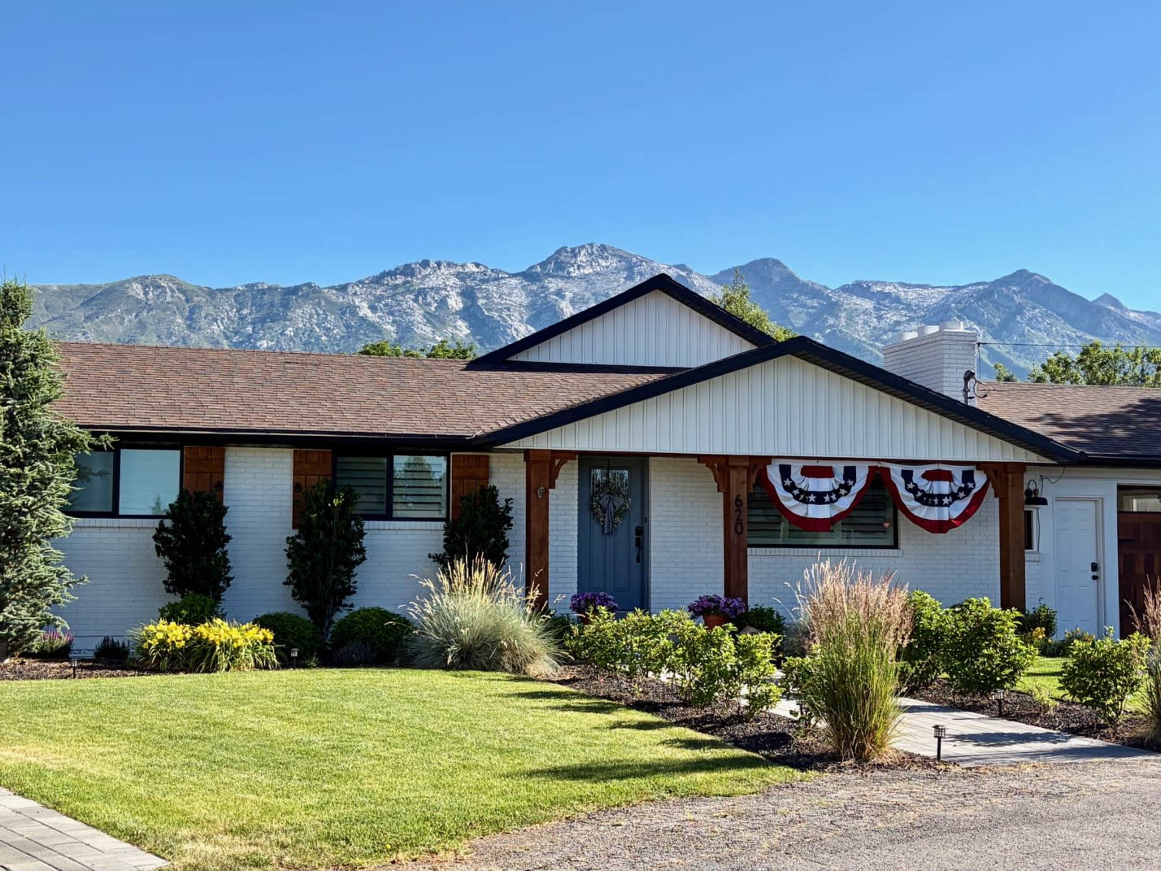 A single-story white house with decorative bunting and a landscaped front yard is situated against a backdrop of mountains.