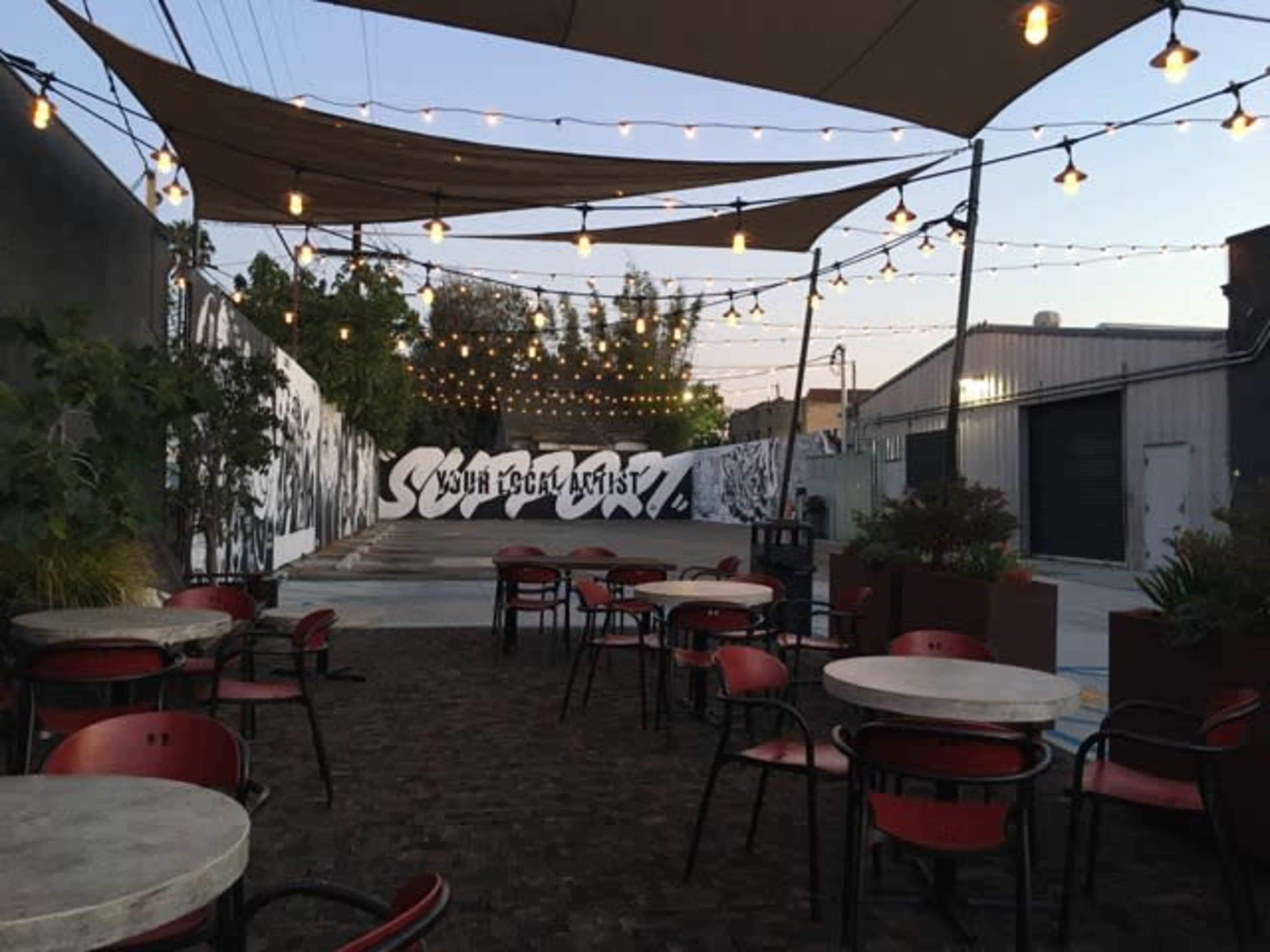 The image shows an outdoor seating area with tables and chairs beneath string lights, adjacent to a wall featuring street art.