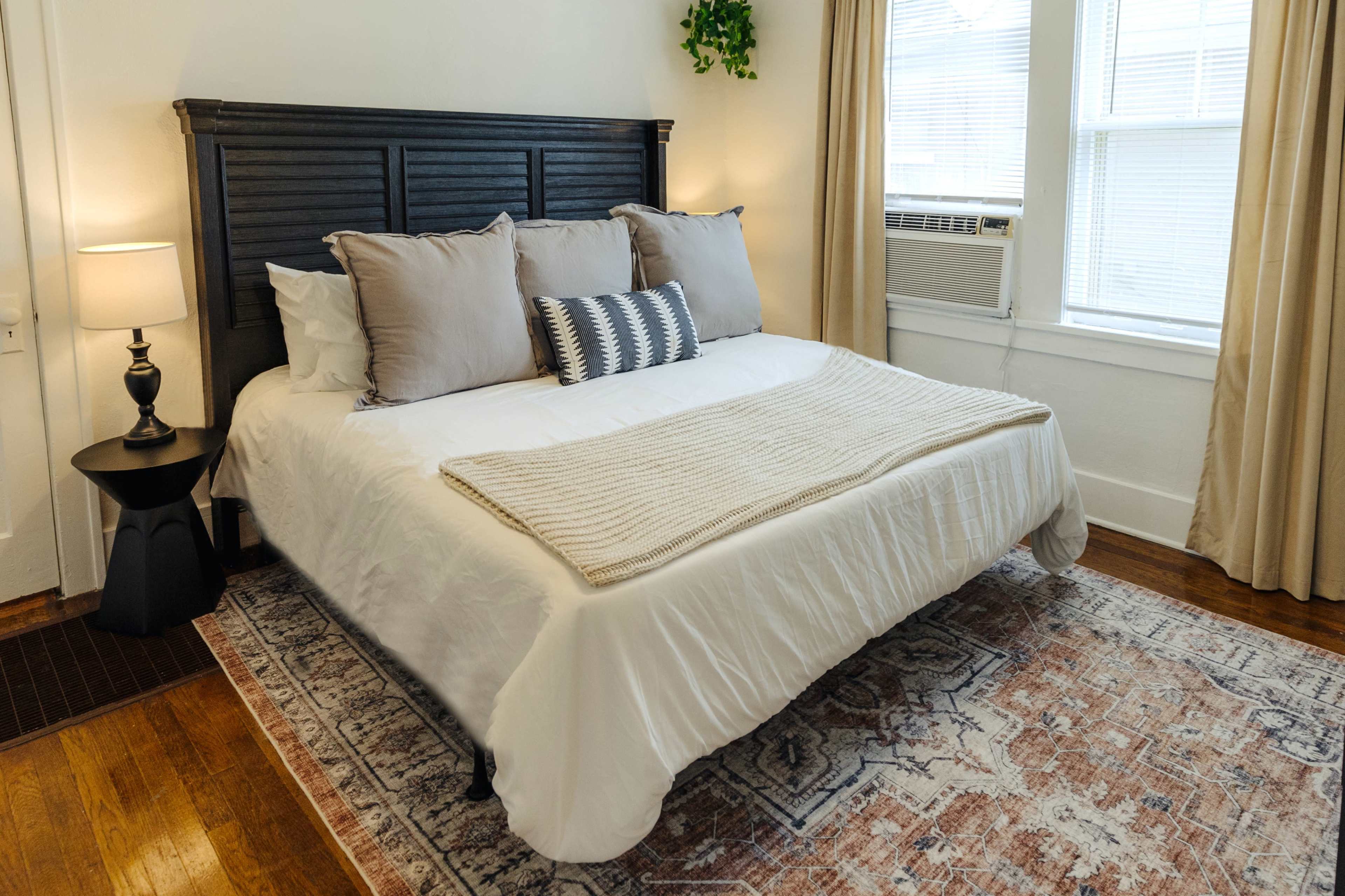 A neatly arranged bedroom features a dark wooden headboard, a queen-sized bed with light bedding, and a patterned area rug on hardwood floors.