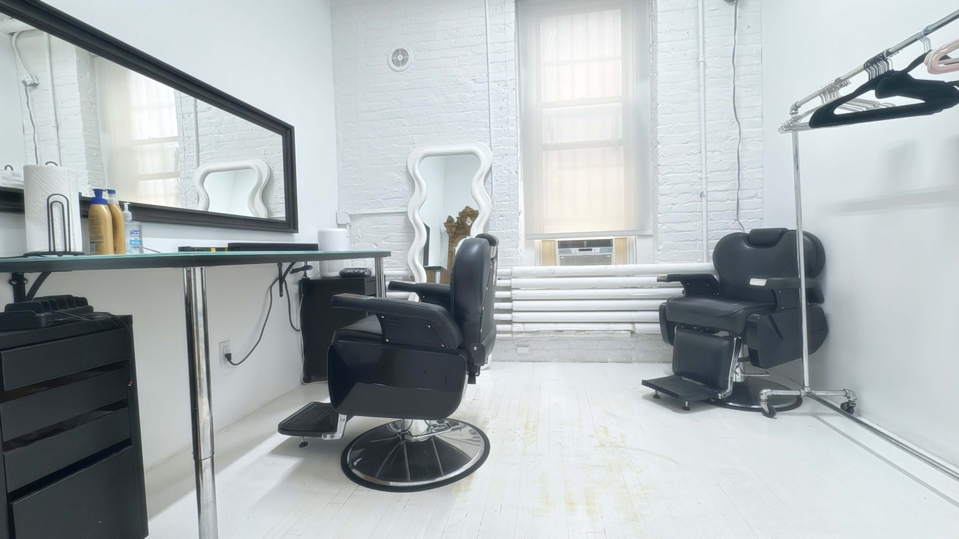 The image shows a bright, minimalist hair salon with two black salon chairs, a large mirror, and a clothing rack on the wall.