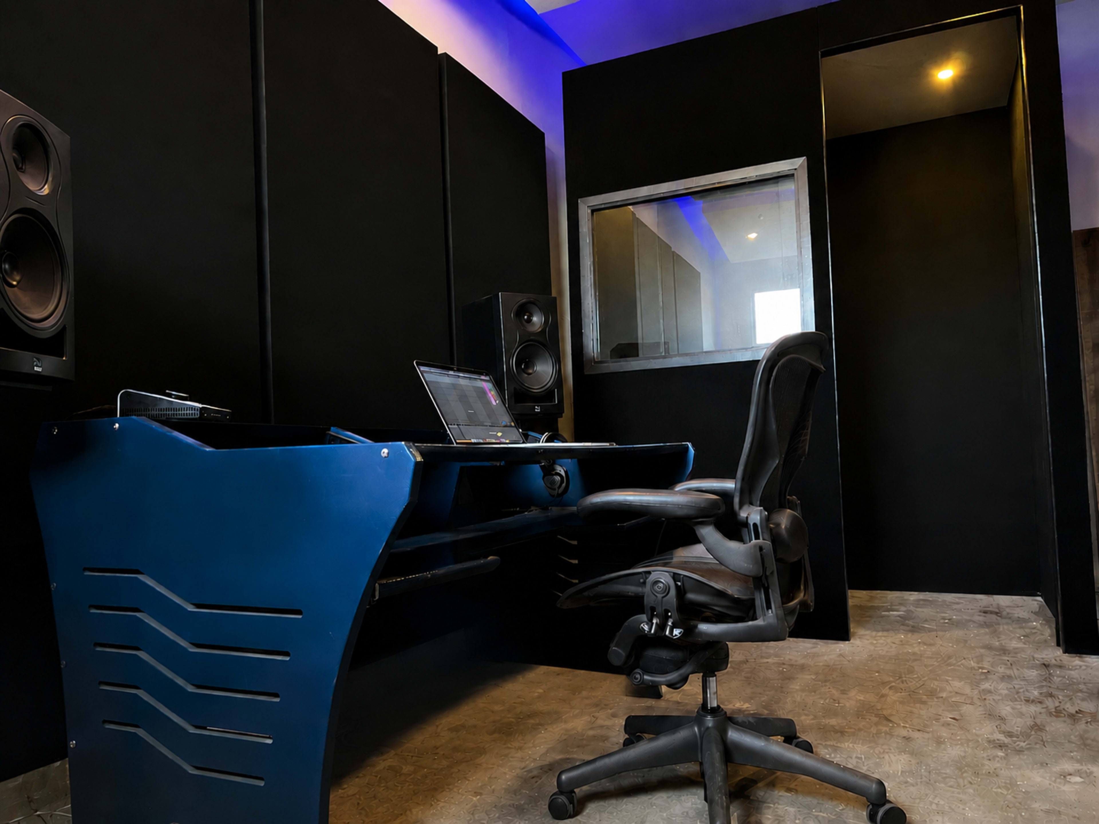 The image shows a modern recording studio with a blue desk, an office chair, and soundproof panels on the walls.