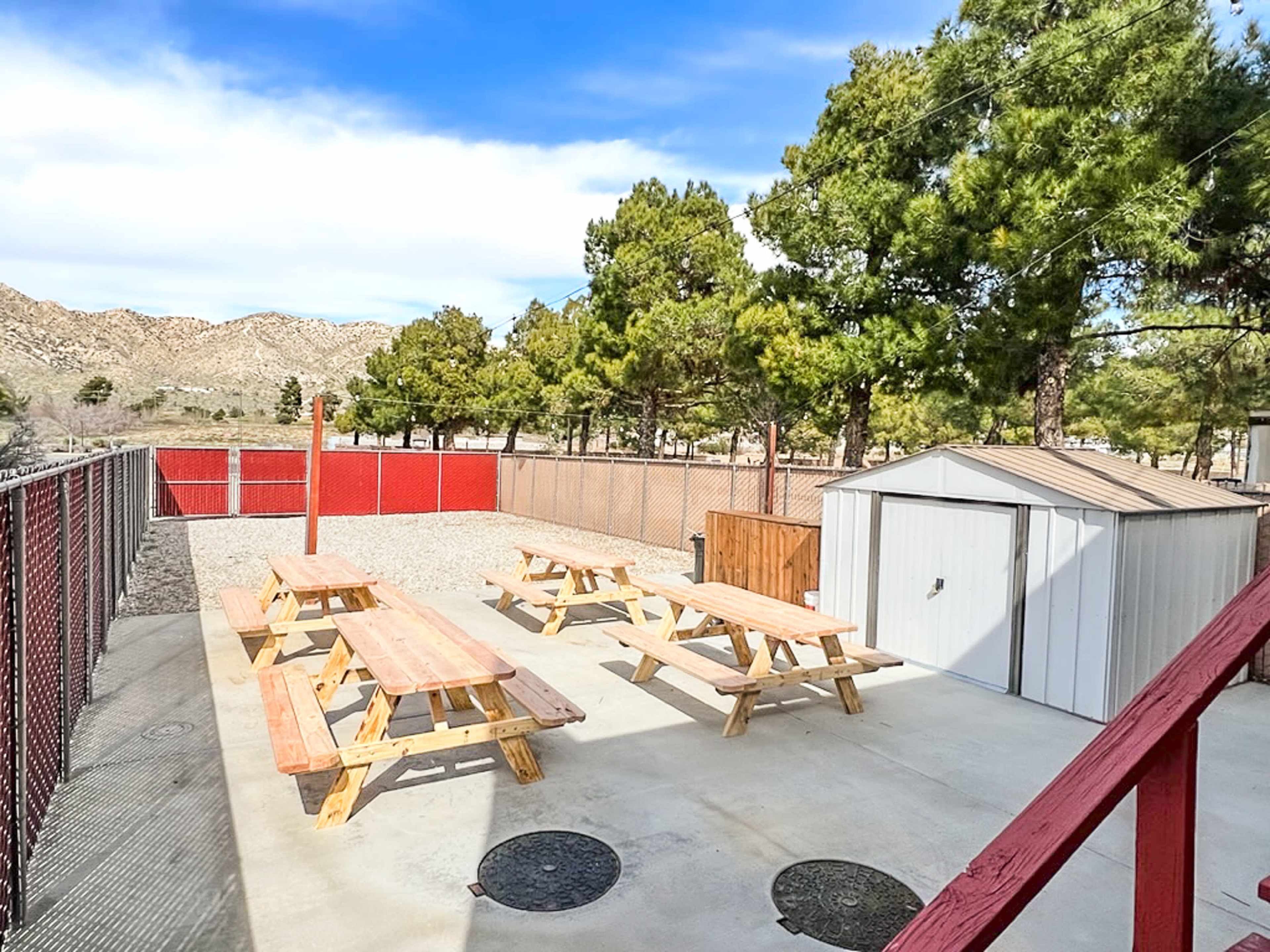 A fenced outdoor area with several picnic tables under trees, adjacent to a small storage shed.