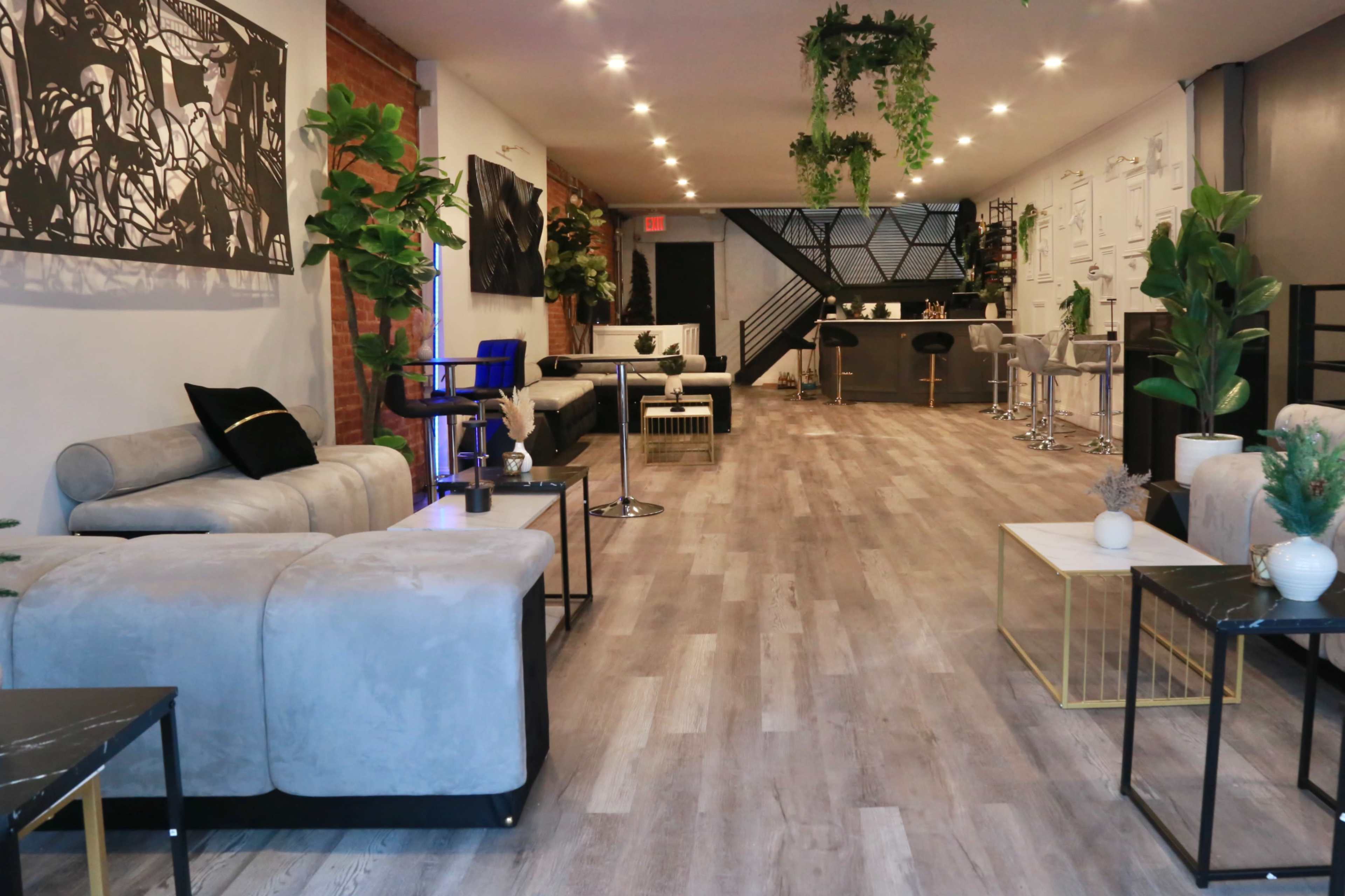 A modern, spacious lounge area with comfortable seating, wooden flooring, and plants, illuminated by overhead lights.