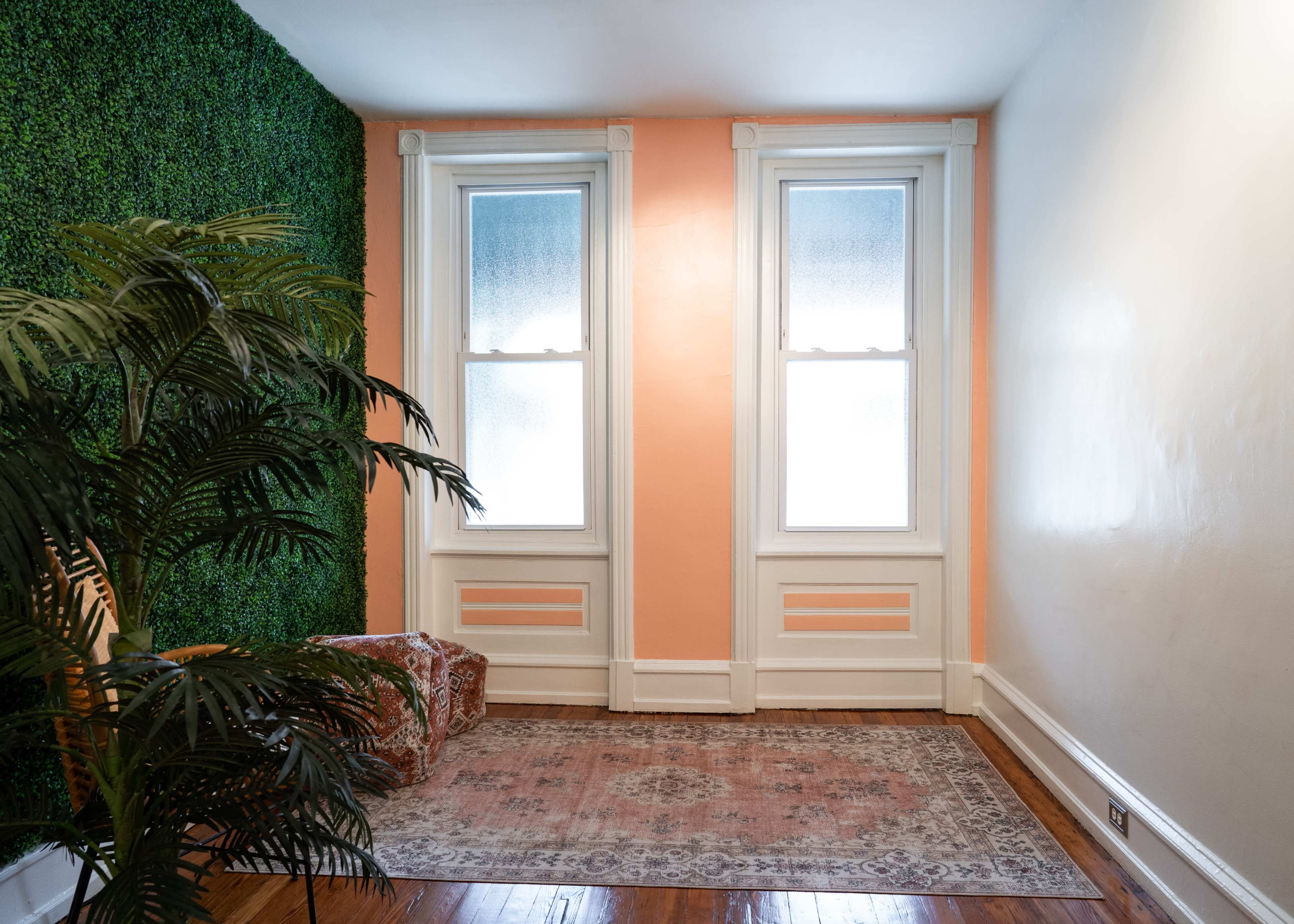 A room with a green wall decoration, two windows, and an orange-painted wall, featuring a rug and a small couch.