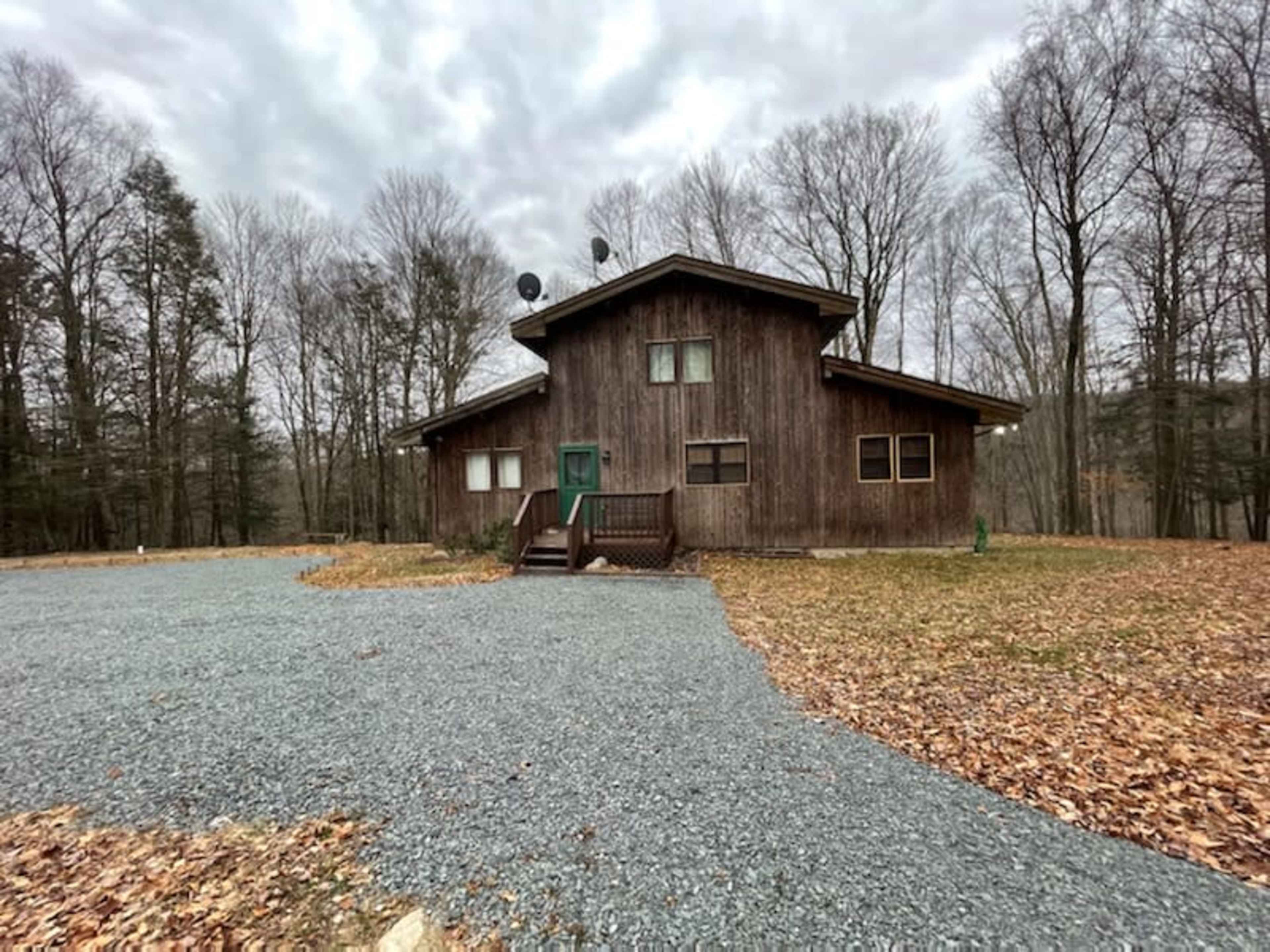 A wooden house with a sloped roof and green door is situated in a gravel-lined clearing surrounded by trees.