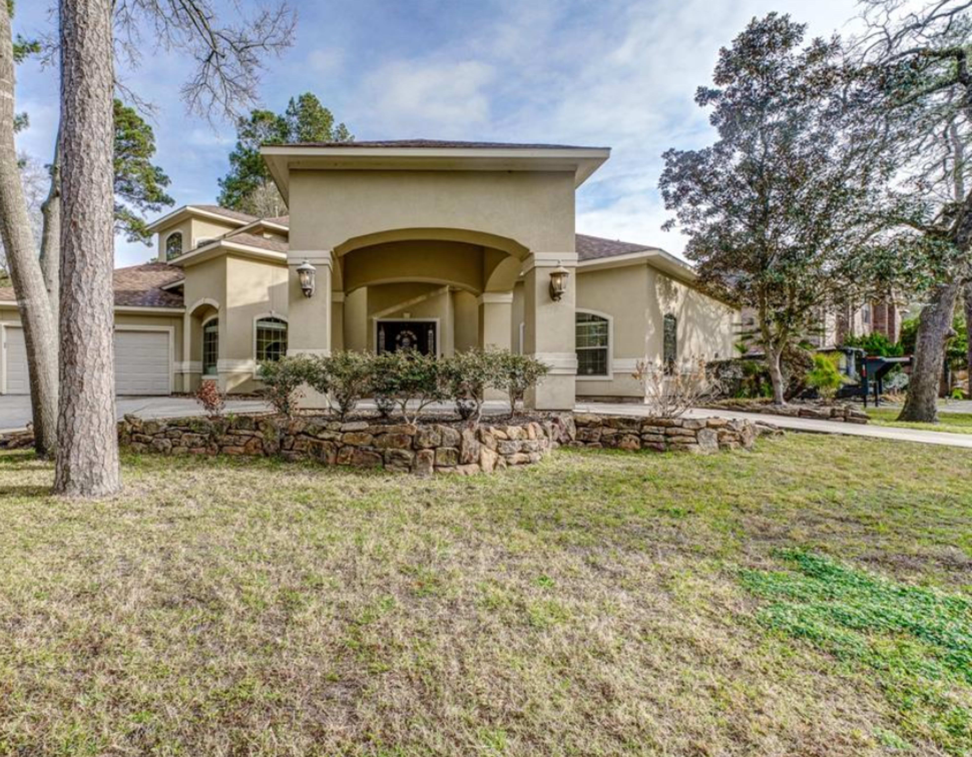 A single-story house with a light-colored exterior, a covered entryway, and a landscaped front yard featuring trees and a stone border.