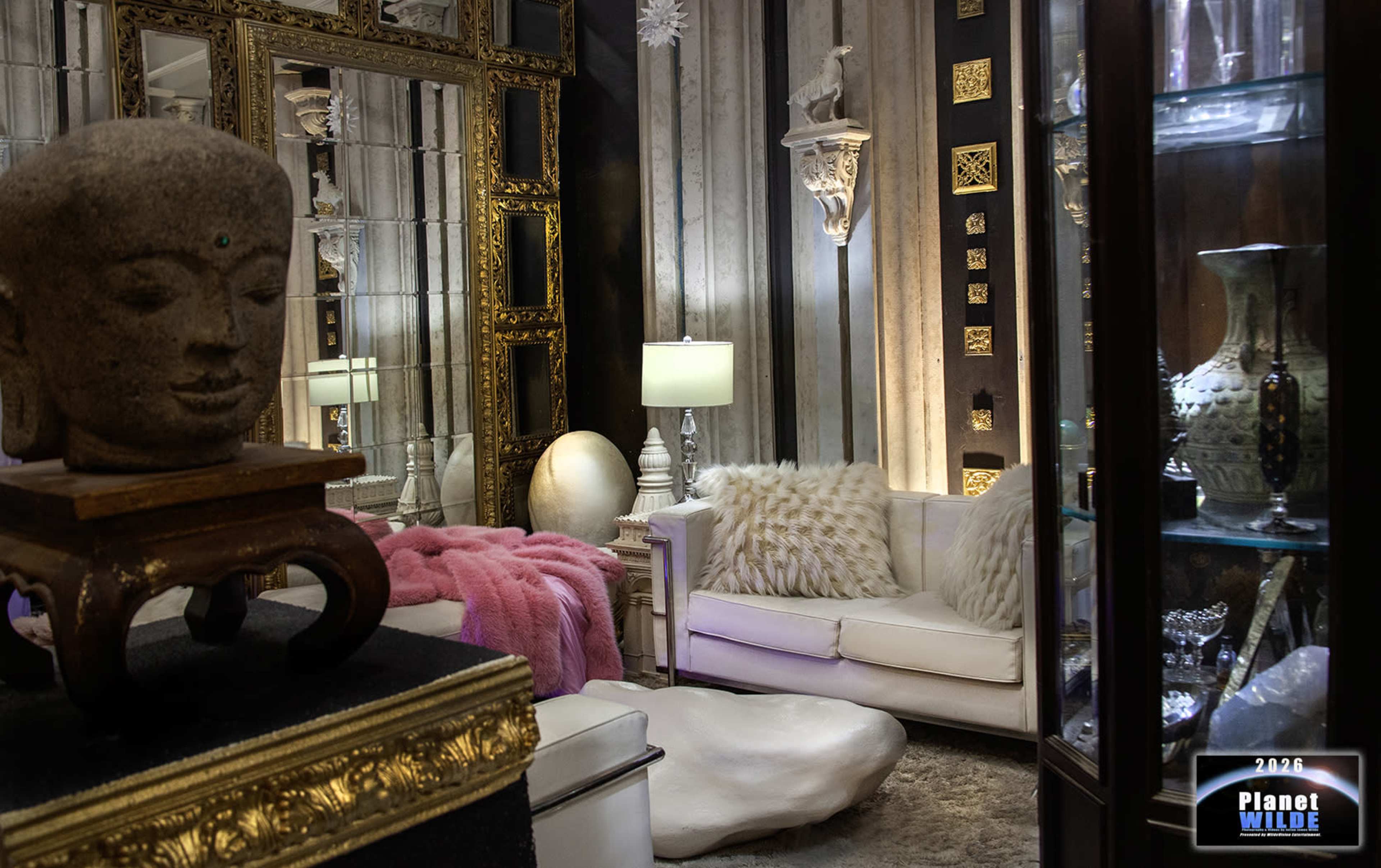 The image shows a stylish interior space featuring a white sofa, decorative mirrors, various sculptures, and display cabinets with intricate items.