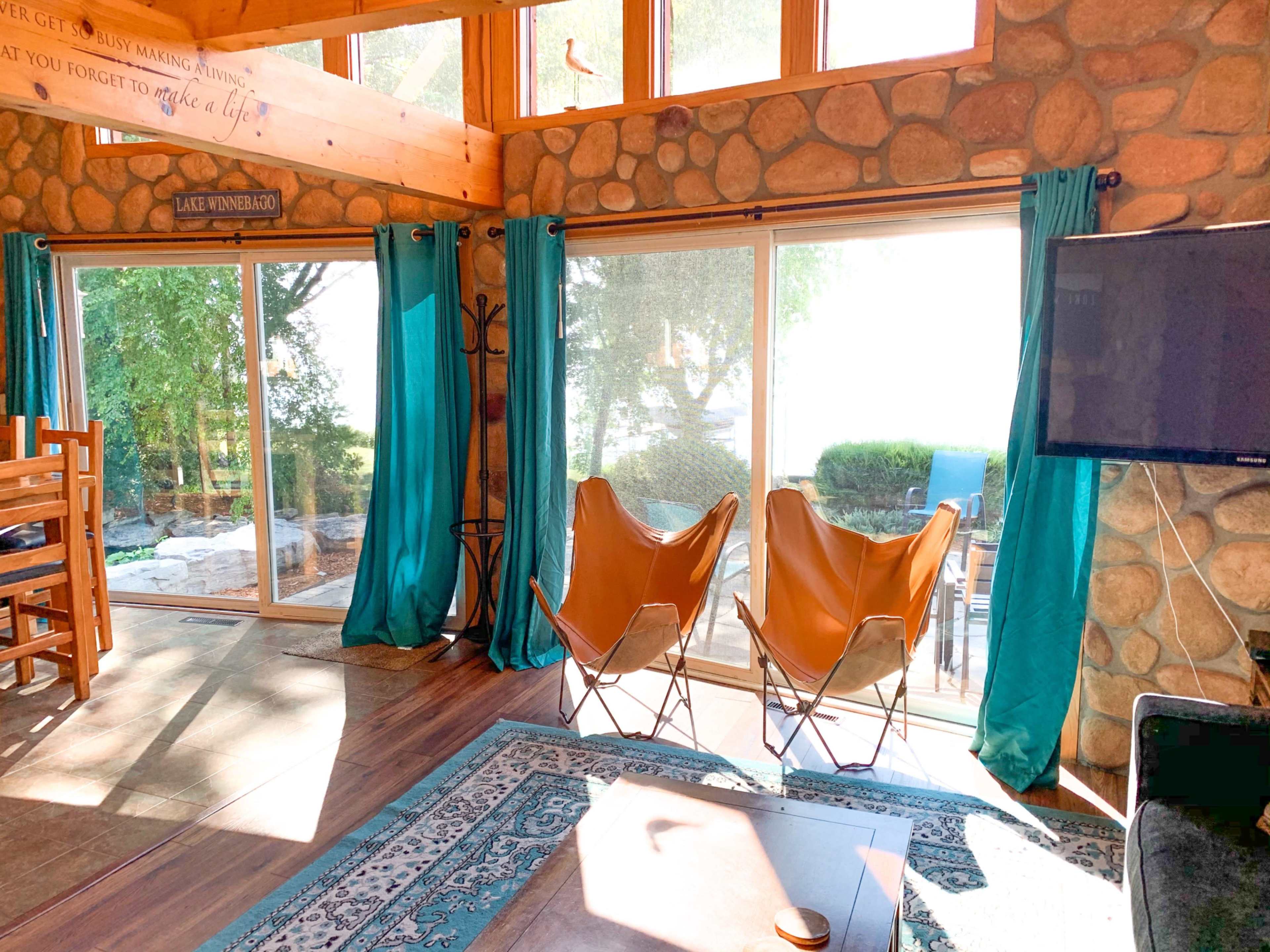 The interior of a cozy room features a stone wall, large windows with sheer curtains, and two orange chairs facing outdoor scenery.