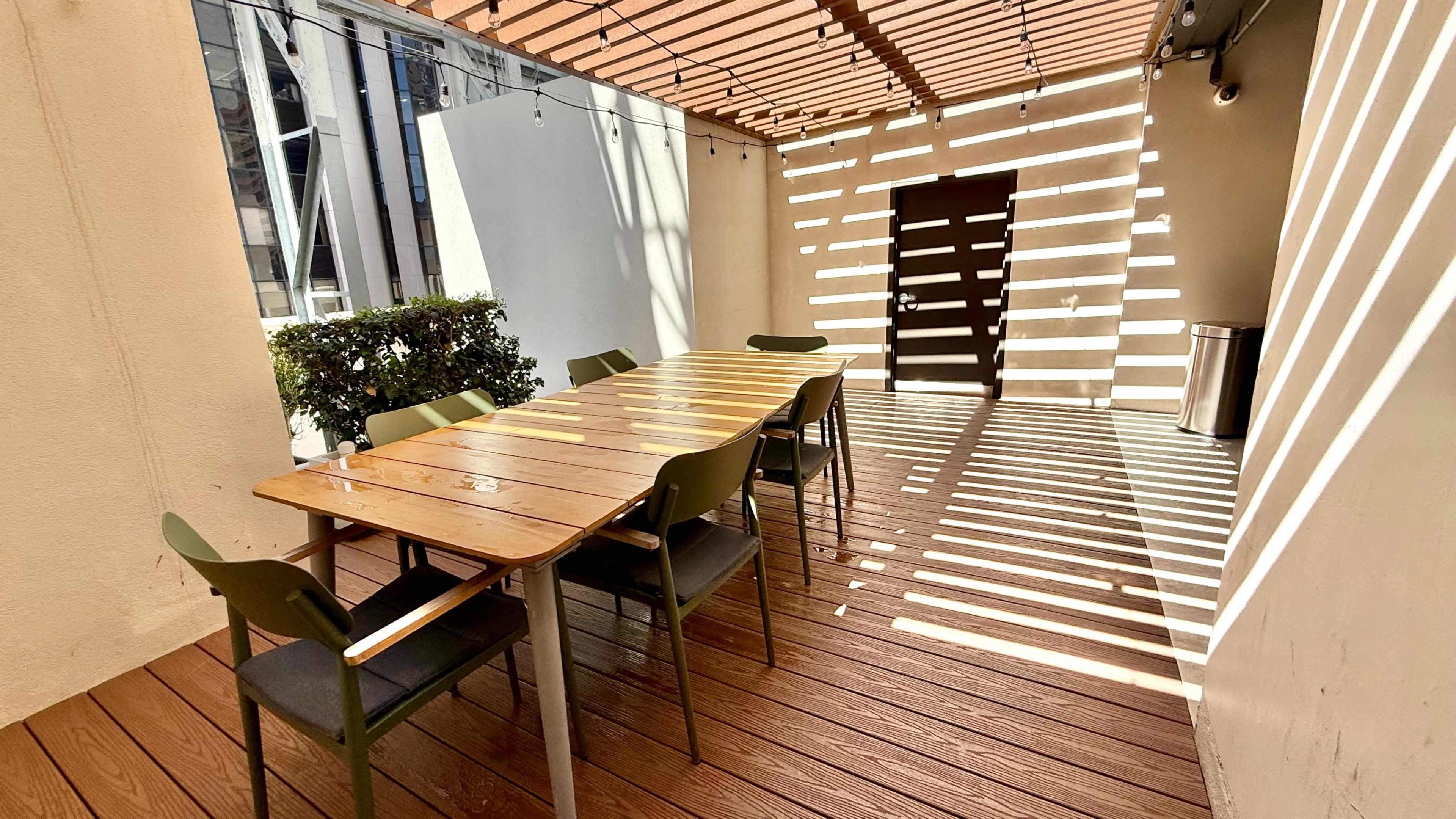 A long wooden table with several chairs is positioned under a slatted roof, casting shadows on the wooden floor in a well-lit outdoor space.