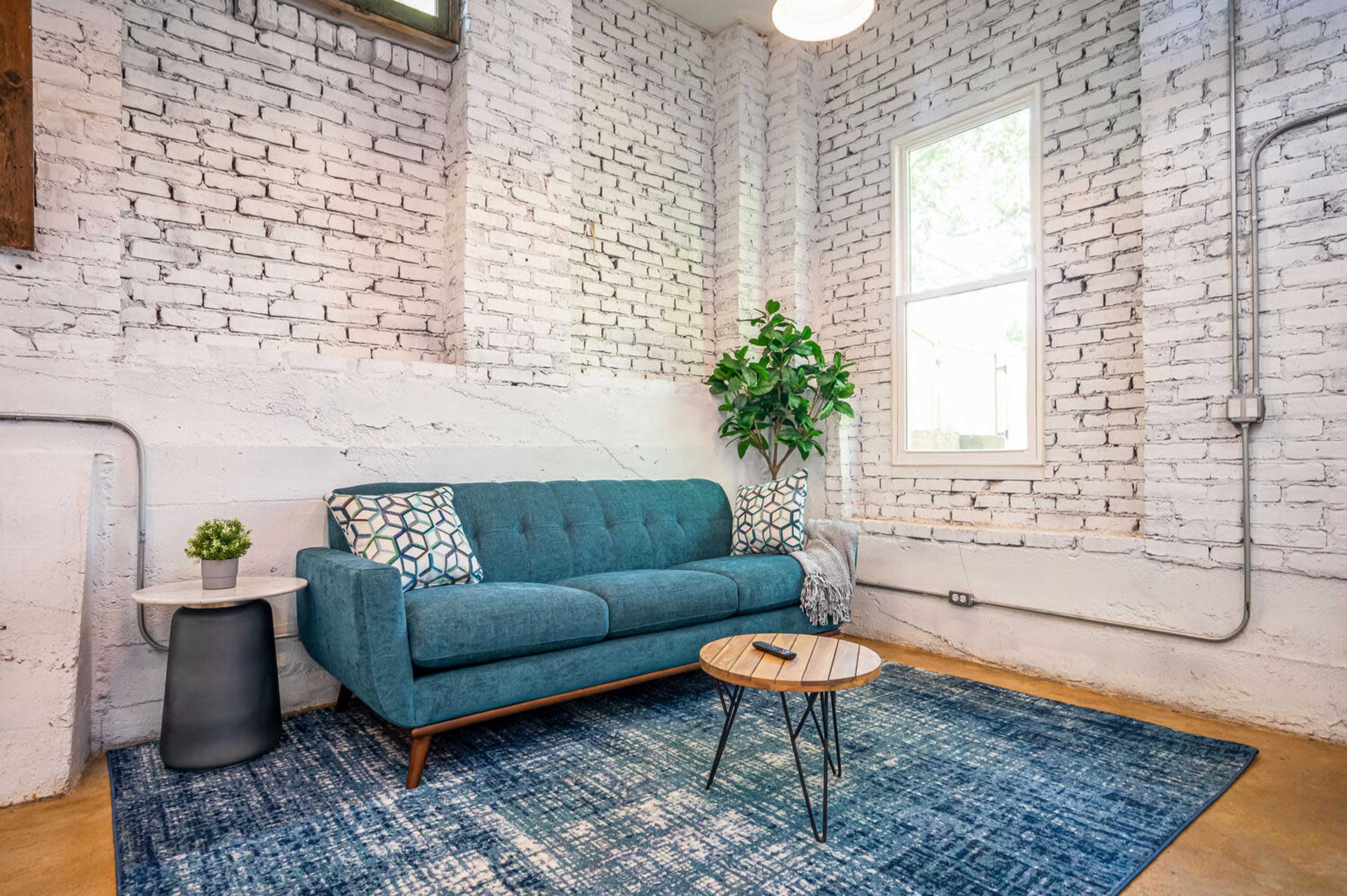 A cozy living room with a teal sofa, a round coffee table, a small plant, and a large area rug, set against a backdrop of white brick walls and a window.