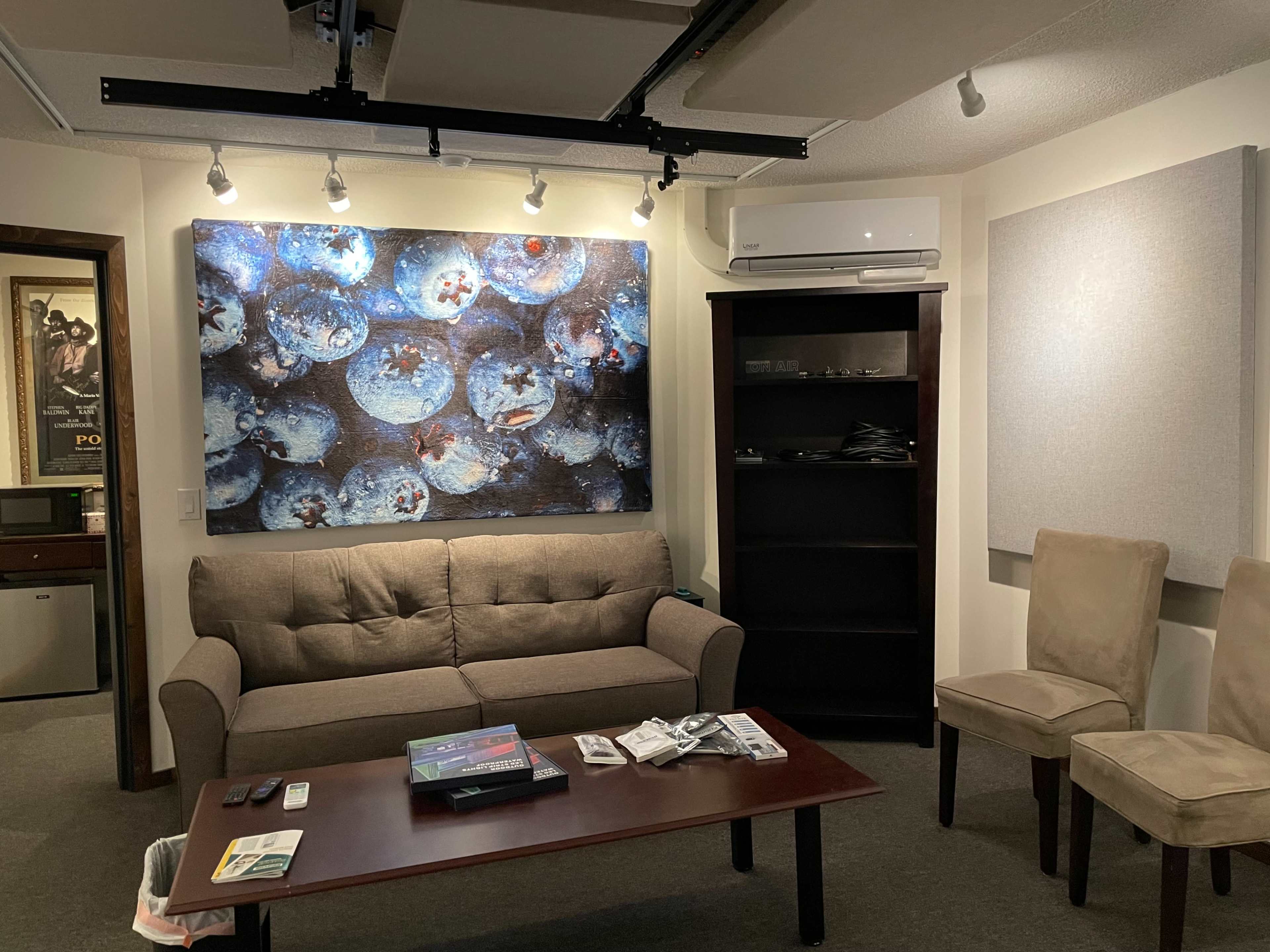 The image shows a cozy room featuring a brown sofa, a wooden coffee table with various items, a bookshelf, two beige chairs, and a large abstract artwork on the wall.
