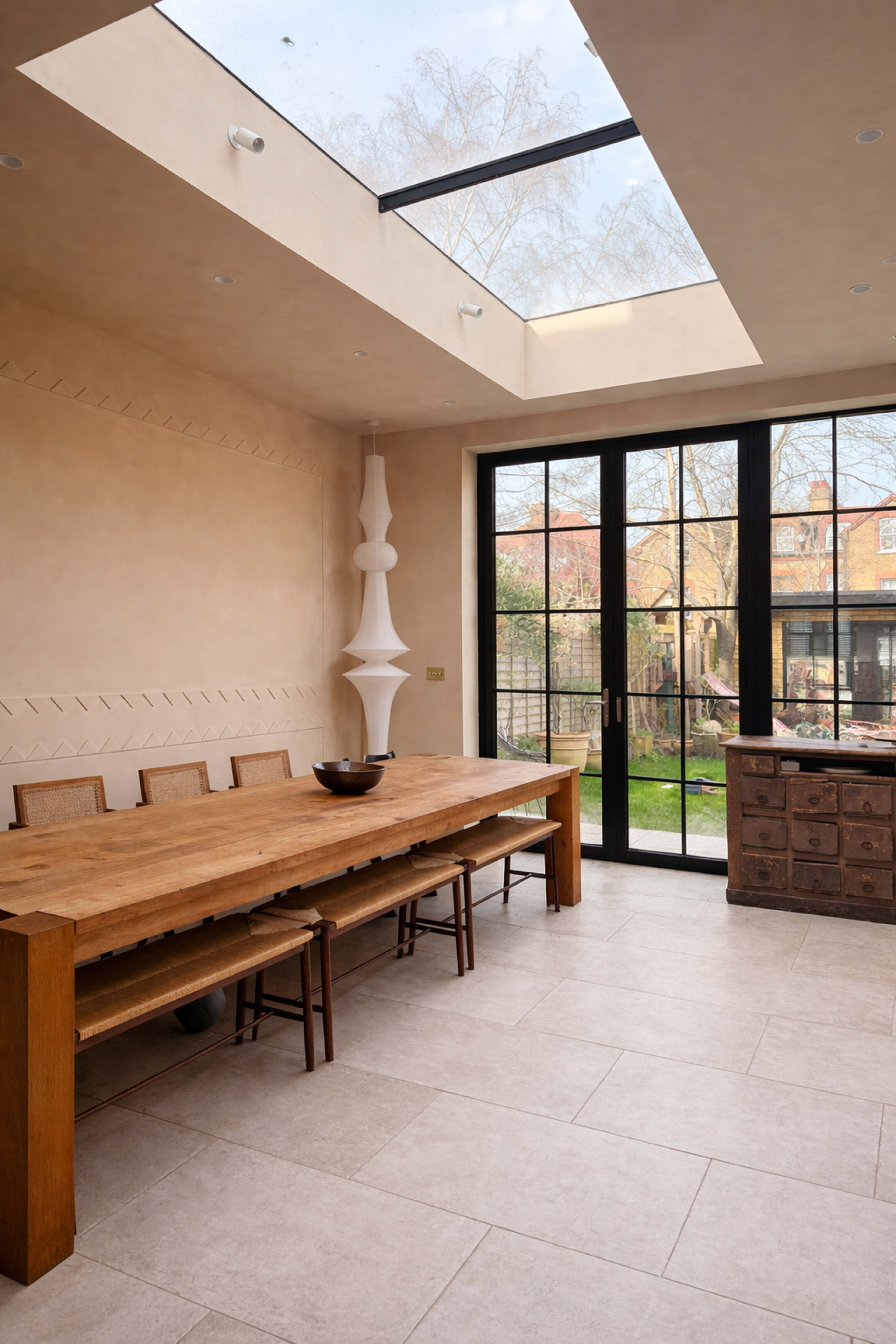 The image shows a spacious dining area with a large wooden table, surrounded by chairs, under a skylight, and featuring a view of a garden through glass doors.