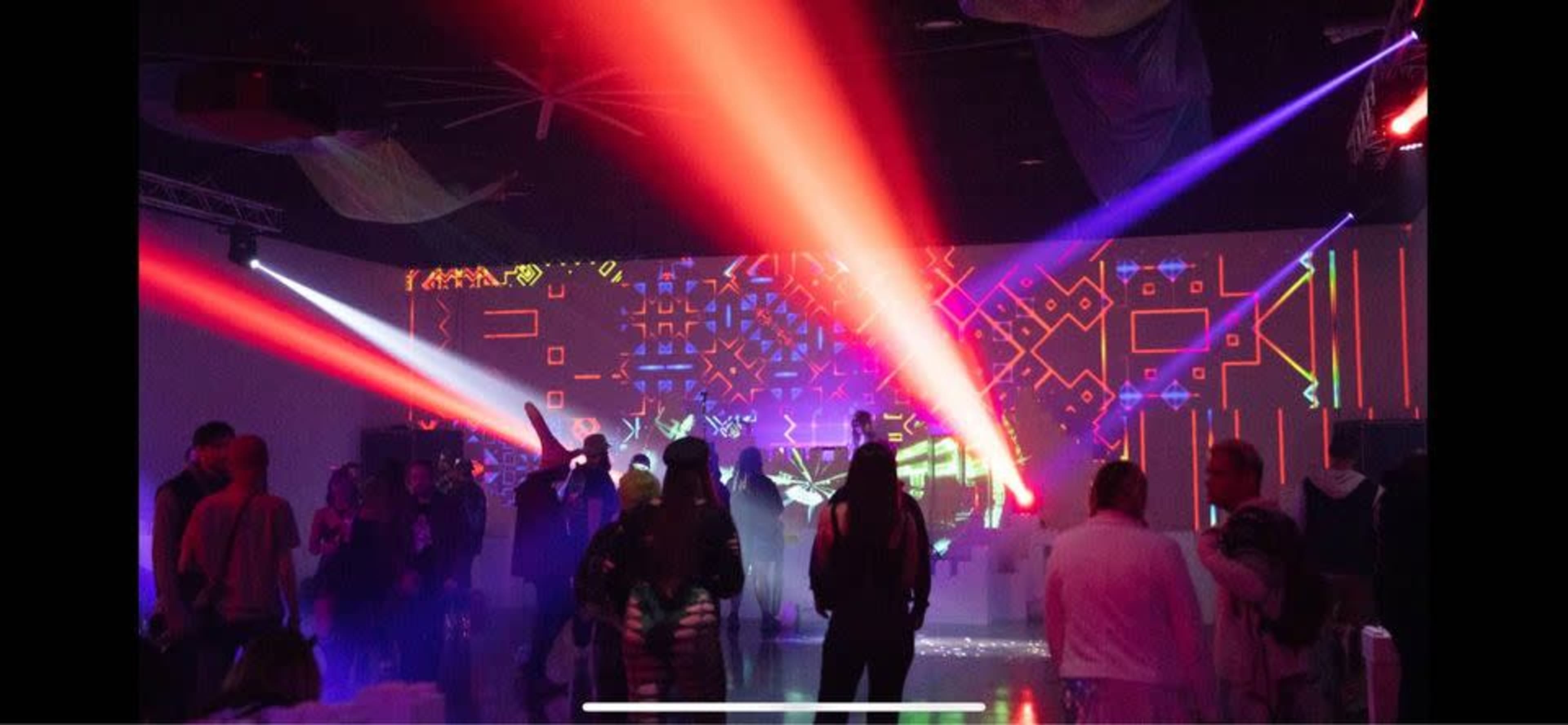 A vibrant indoor party scene features people dancing under colorful laser lights and projected visuals on the walls.