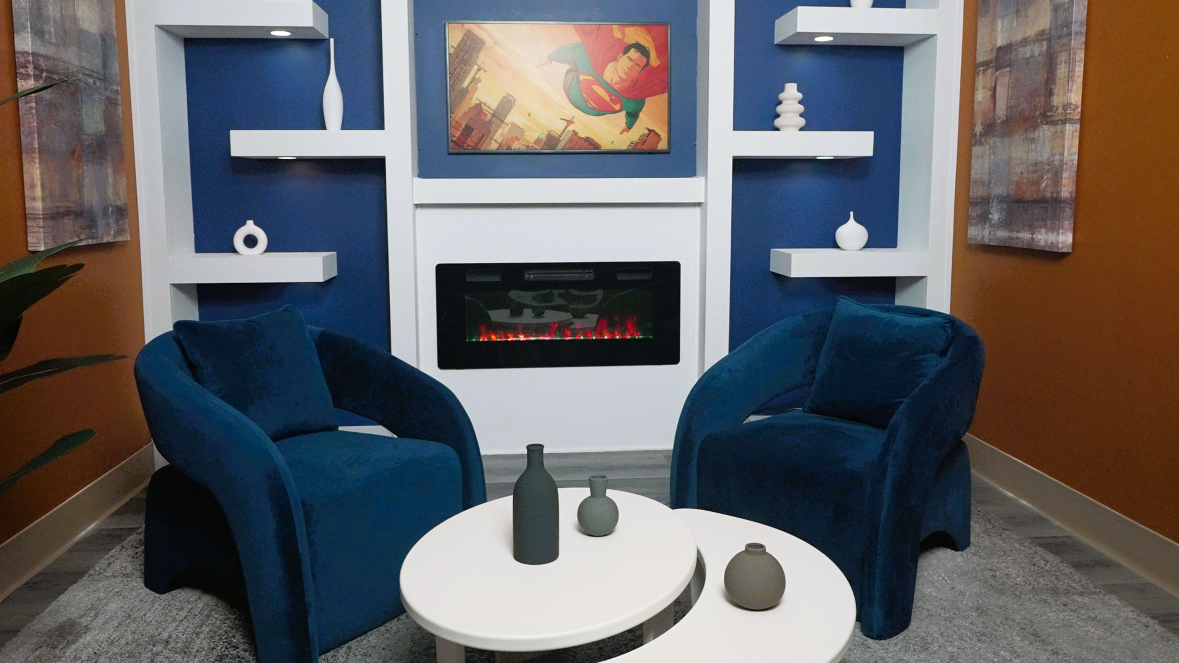 The image shows a modern sitting area featuring two blue chairs, a round white coffee table, and a wall-mounted electric fireplace beneath a framed artwork.