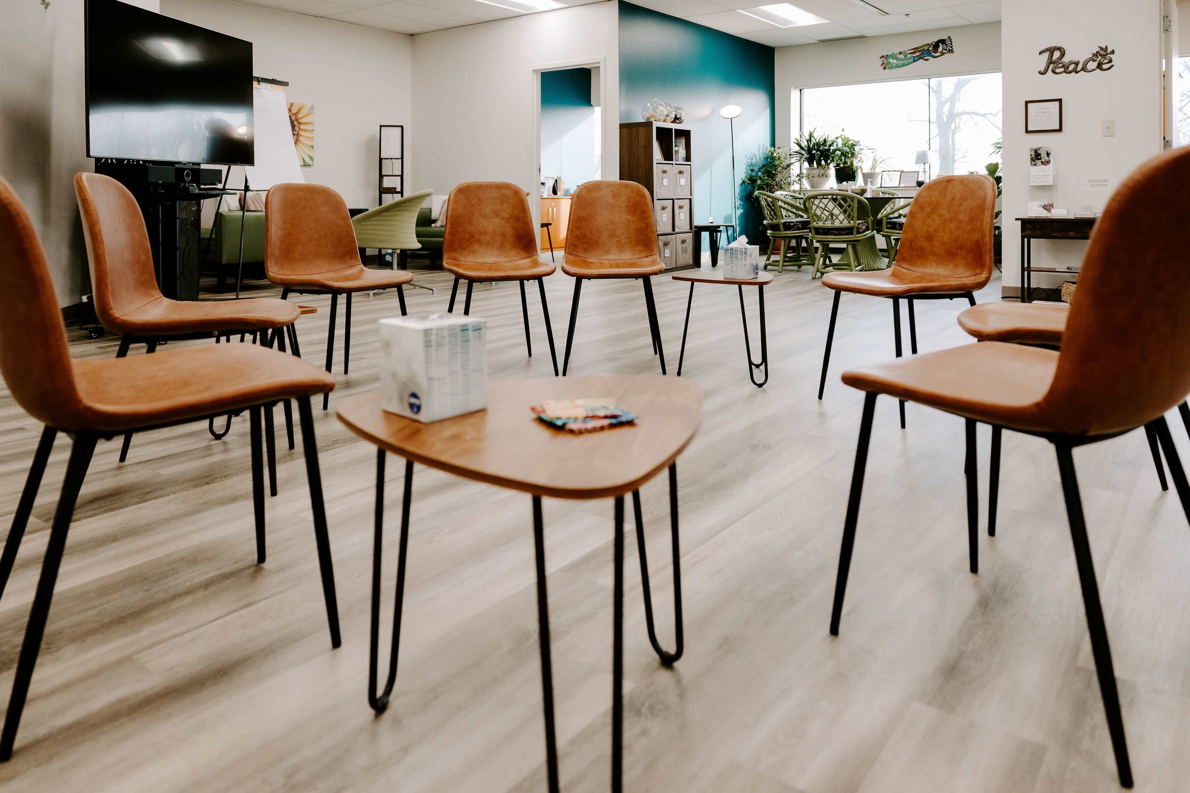 The image shows an arrangement of brown chairs around a small wooden table in a well-lit room with plants and decorative elements in the background.