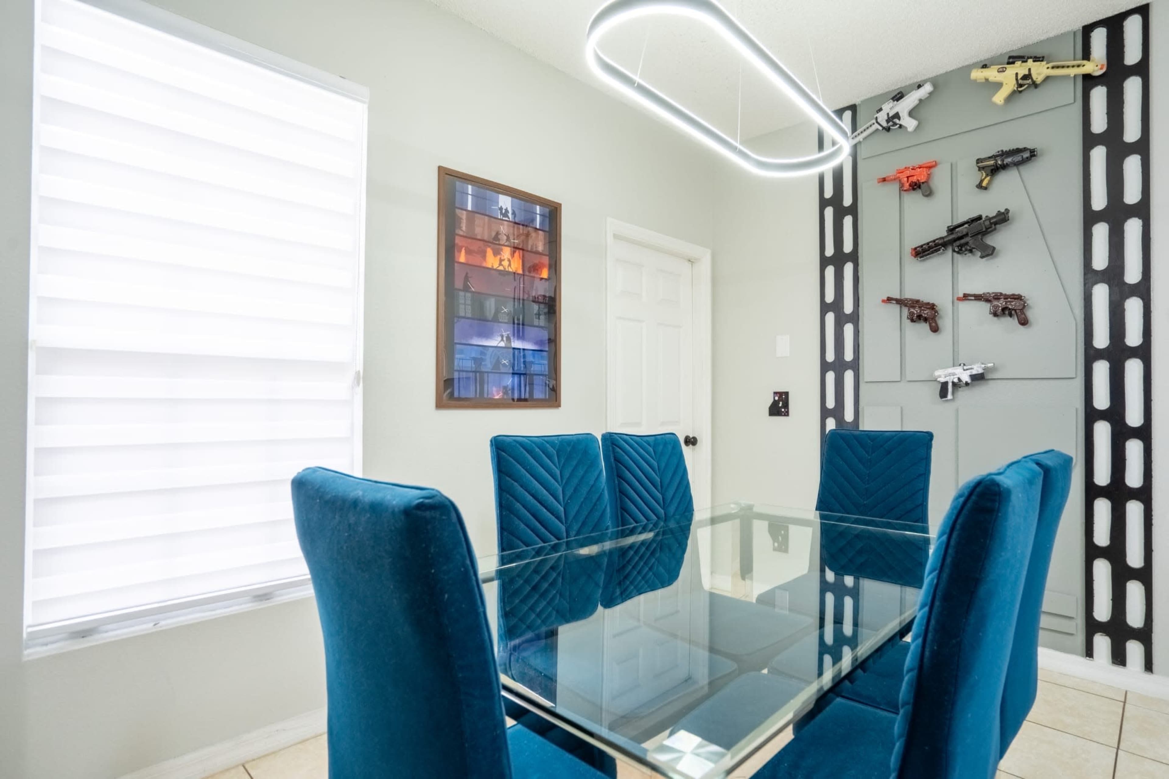 A modern dining area features a glass table surrounded by blue upholstered chairs, with a wall displaying replica firearms and a framed artwork.