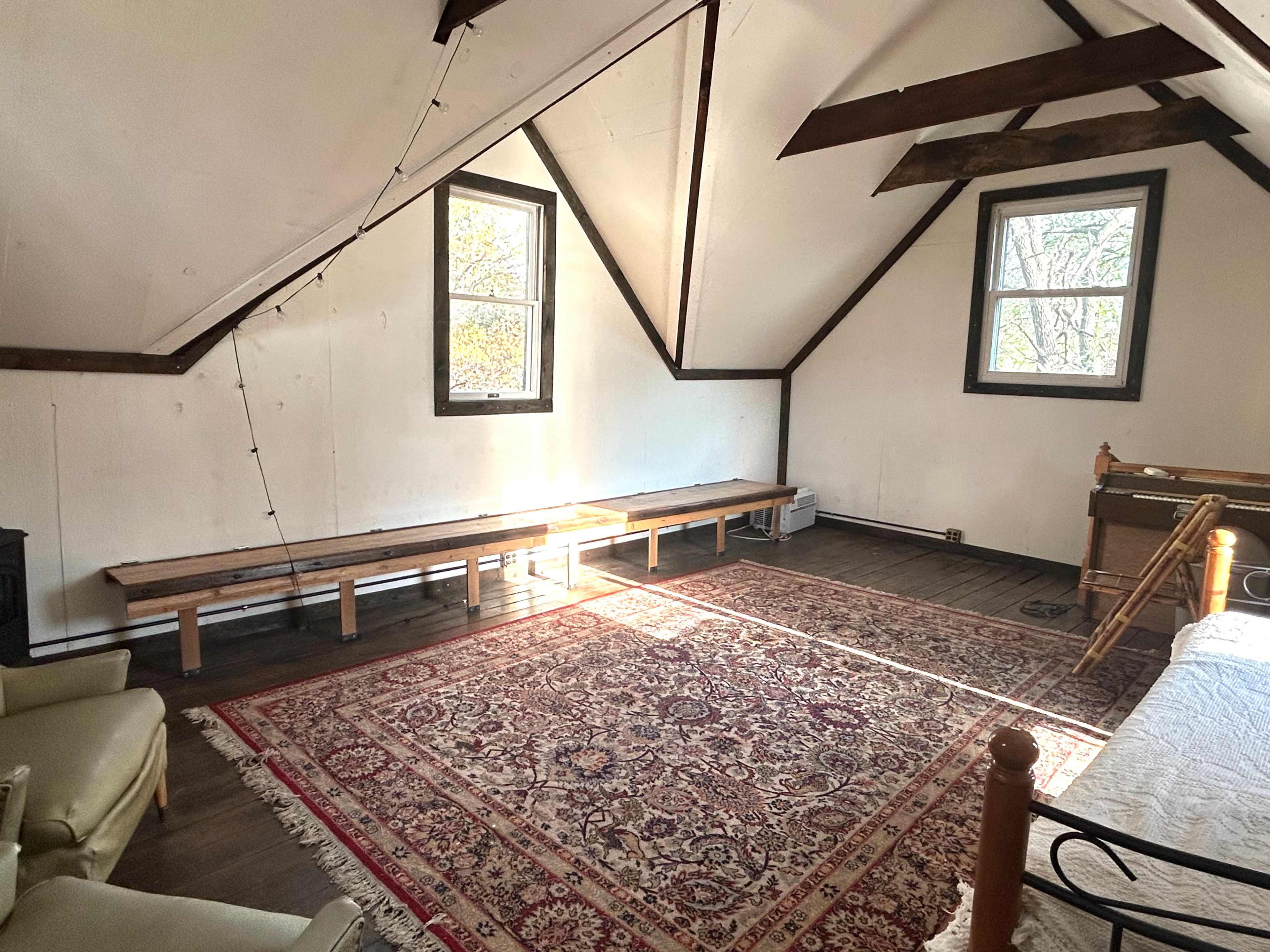 The image shows a spacious, sparsely furnished room with wooden beams, two windows, and a large area rug on the floor.