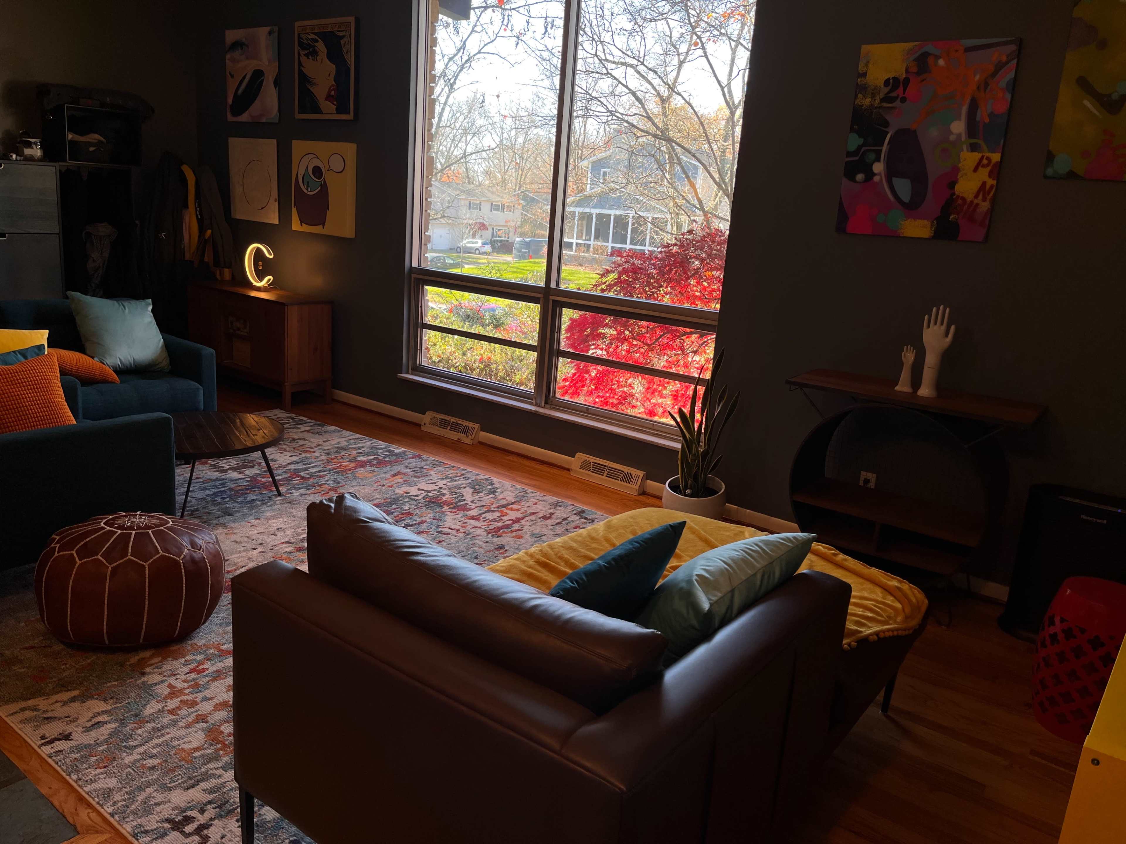 A cozy living room with a sofa, colorful cushions, artwork on the walls, and a large window overlooking a yard with trees in autumn colors.