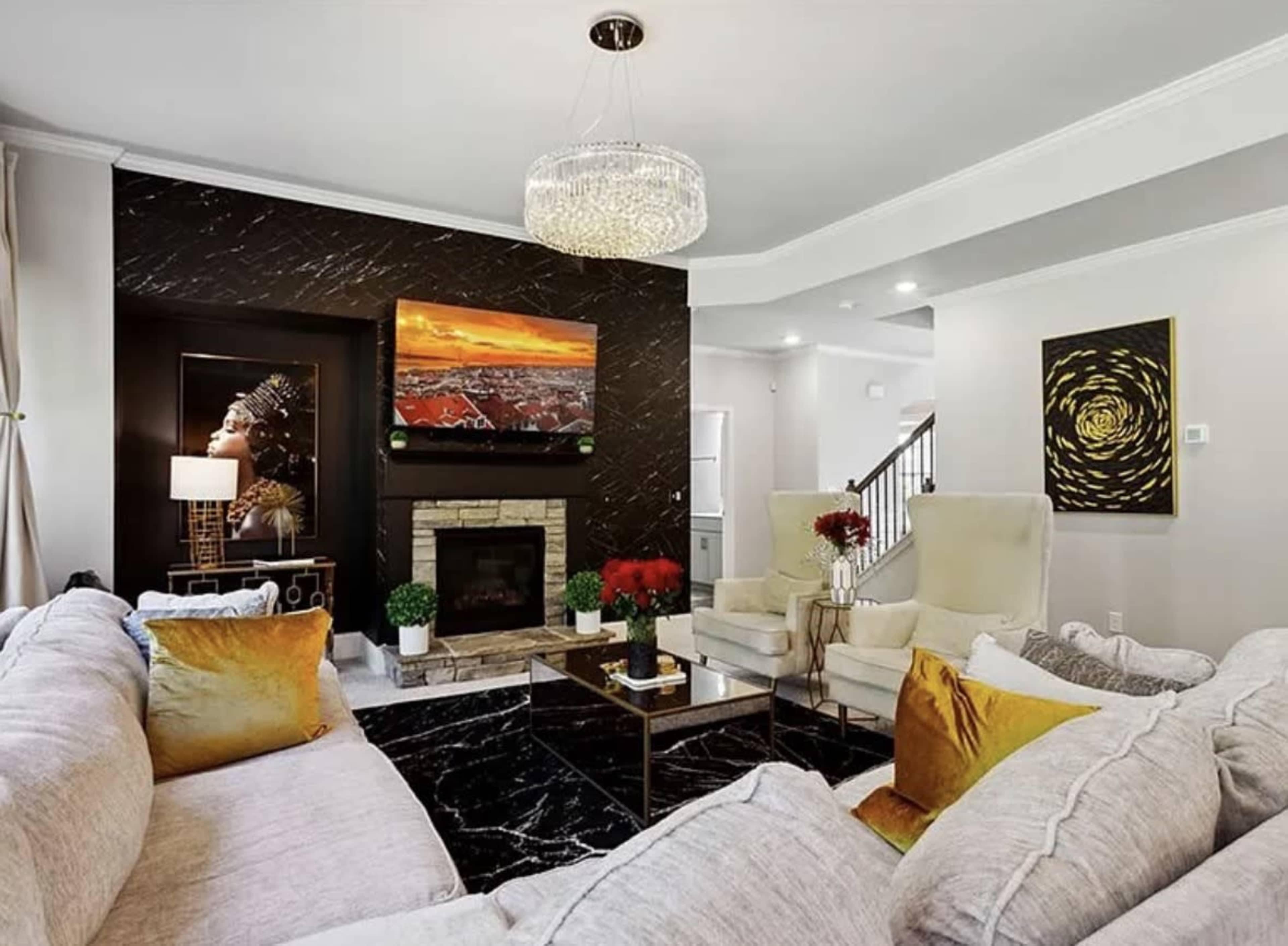 The image shows a modern living room featuring a plush gray sofa, two light-colored armchairs, a marble coffee table, and a fireplace, with artwork and decorative plants on display.