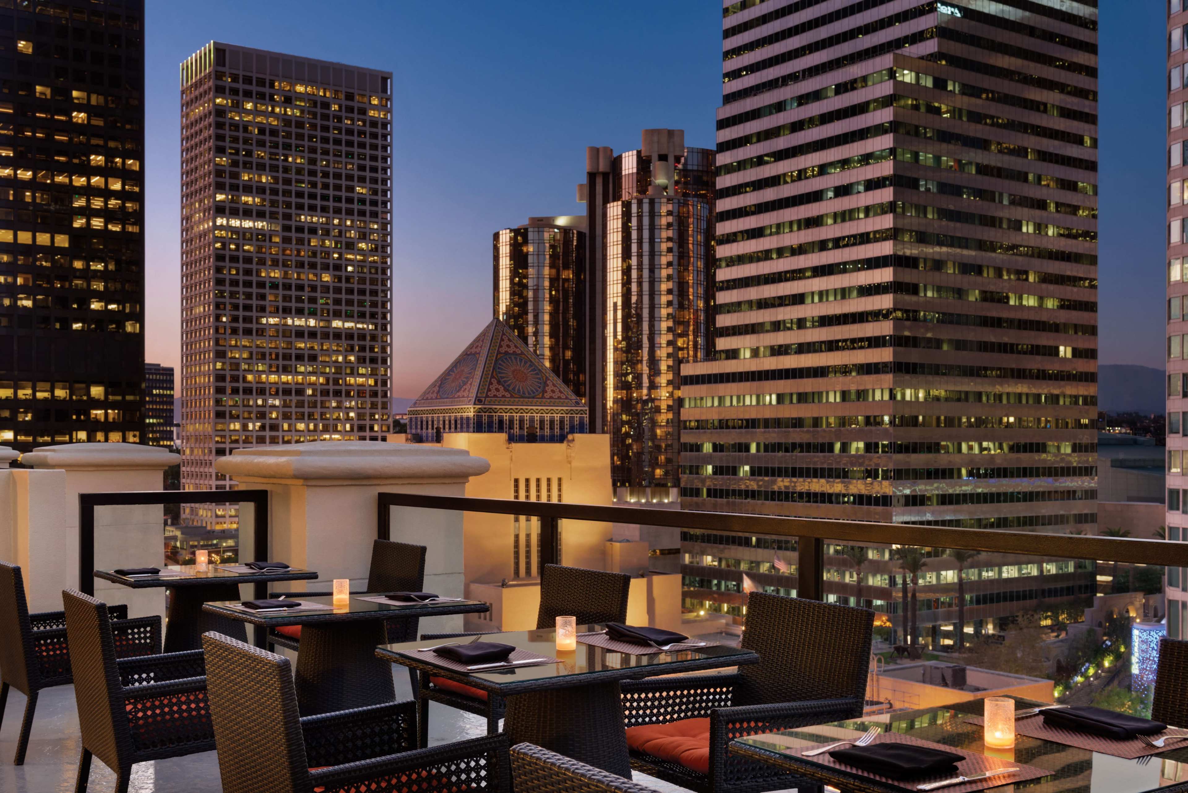 The image shows a rooftop dining area with tables overlooking a city skyline at dusk, featuring tall buildings and lingering twilight colors in the sky.