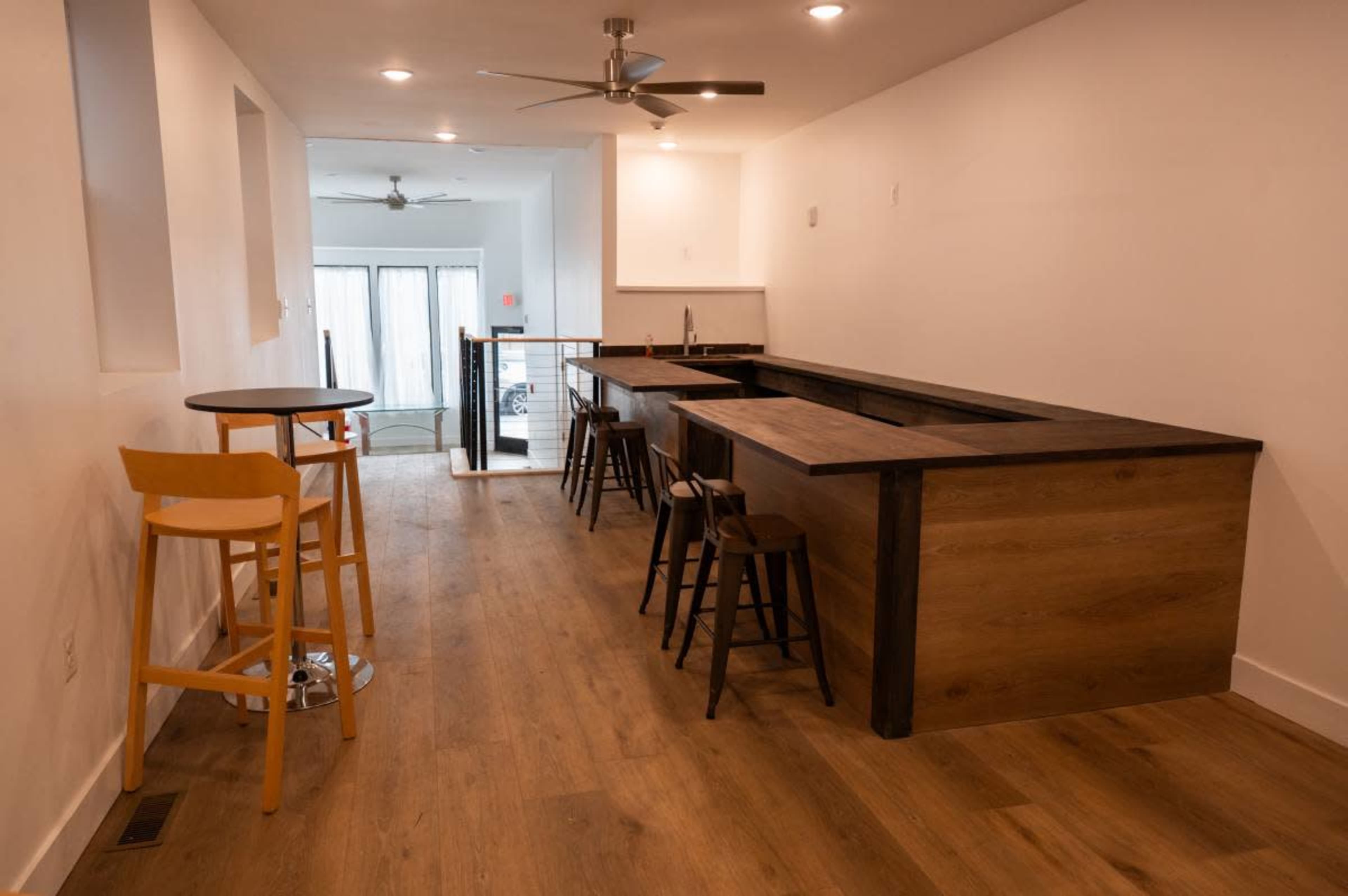 The image shows a modern, elongated room featuring a kitchen bar with wooden countertops, high chairs, and a sitting area beside it, illuminated by overhead lights.
