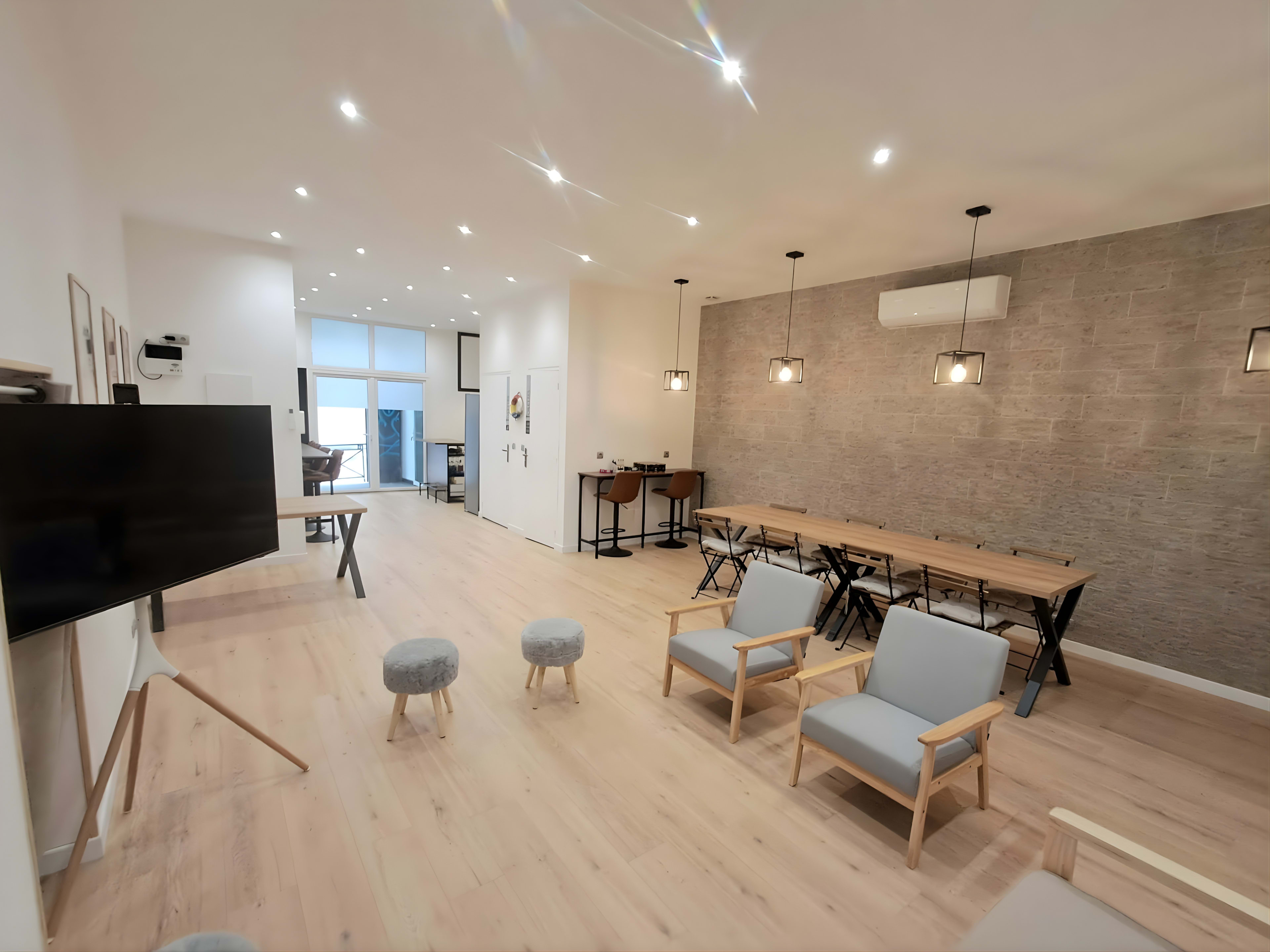 The image shows a modern indoor space featuring wooden floors, a seating area with chairs and stools, a large table, and a stone accent wall.